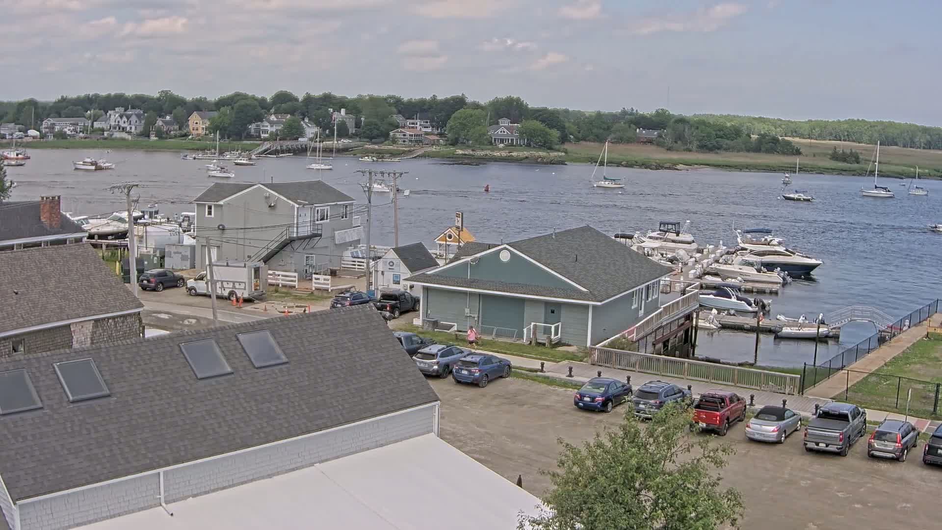 A waterfront scene on a partly cloudy day shows a marina filled with boats, a parking lot with cars, and houses lining a river.