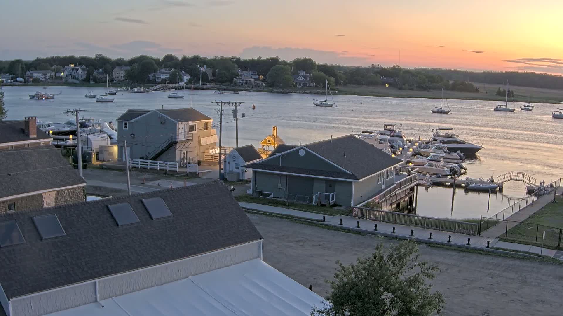 An aerial view at sunset shows a waterfront area with several buildings, boats docked at a marina, and other boats on a calm river, surrounded by trees and houses.