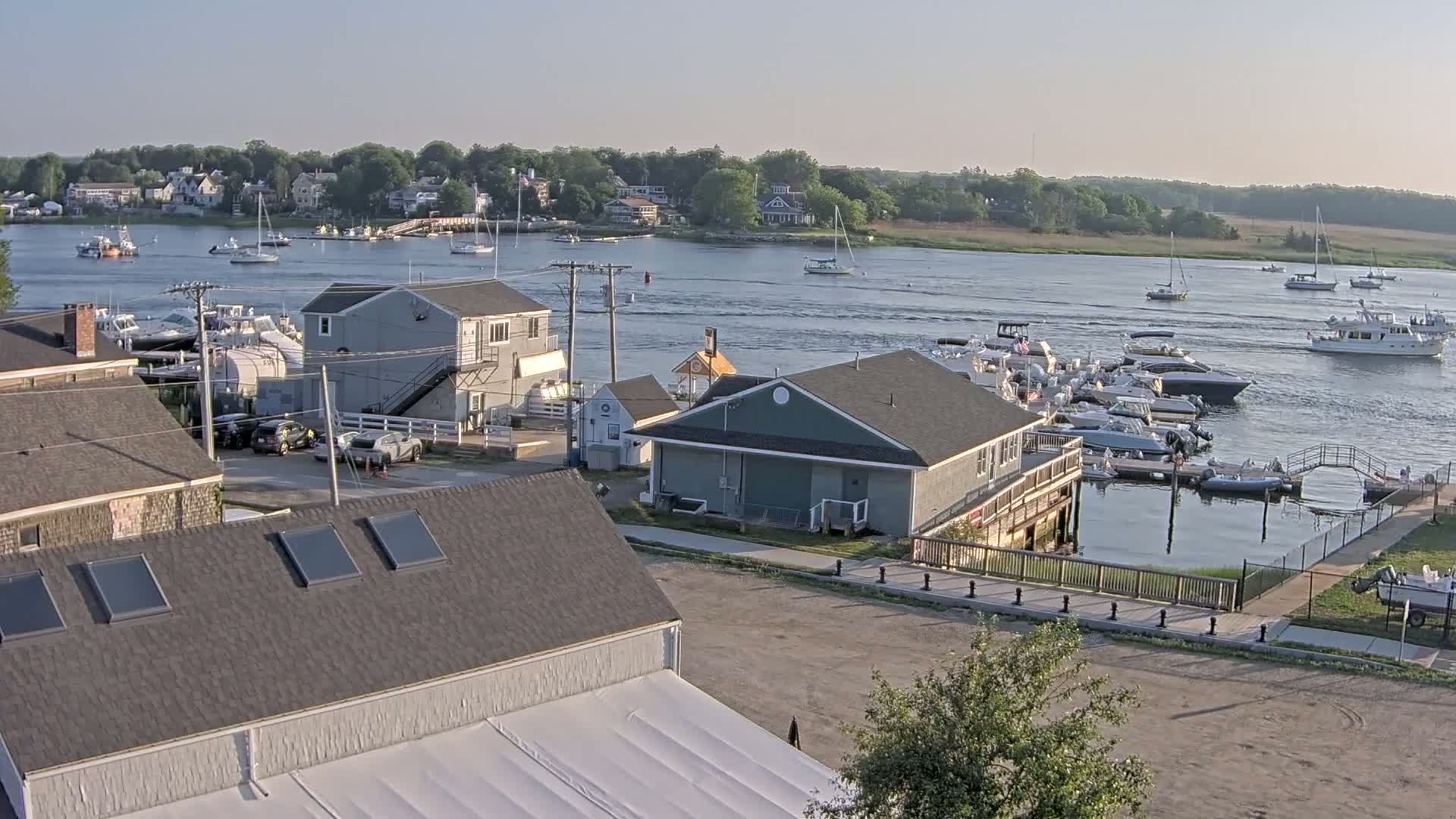 A waterfront town on a calm, sunny day features several boats docked at a marina and others sailing on the river, with houses and buildings lining the shoreline.