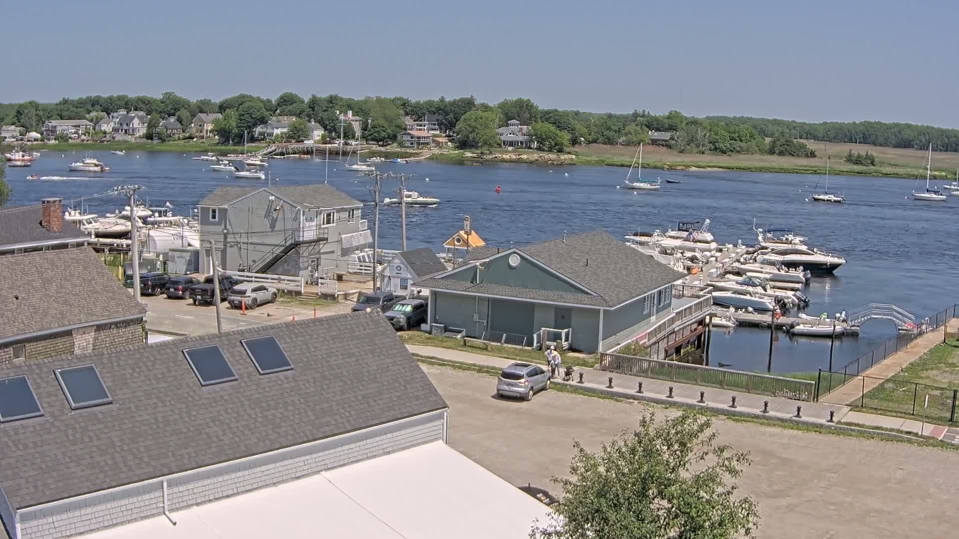 A sunny day reveals a waterfront scene with numerous boats docked at a marina, houses and buildings lining the shore, and several cars parked nearby.