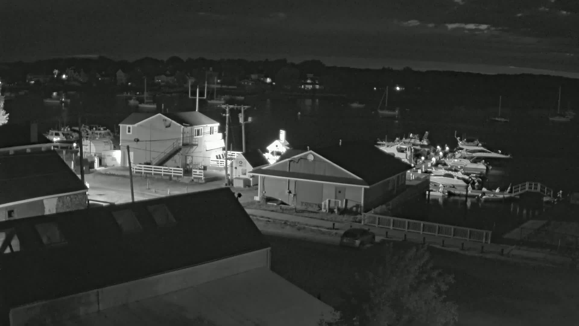 A nighttime view of a harbor with several boats docked, buildings, and a few cars parked along a roadway under a partly cloudy sky.