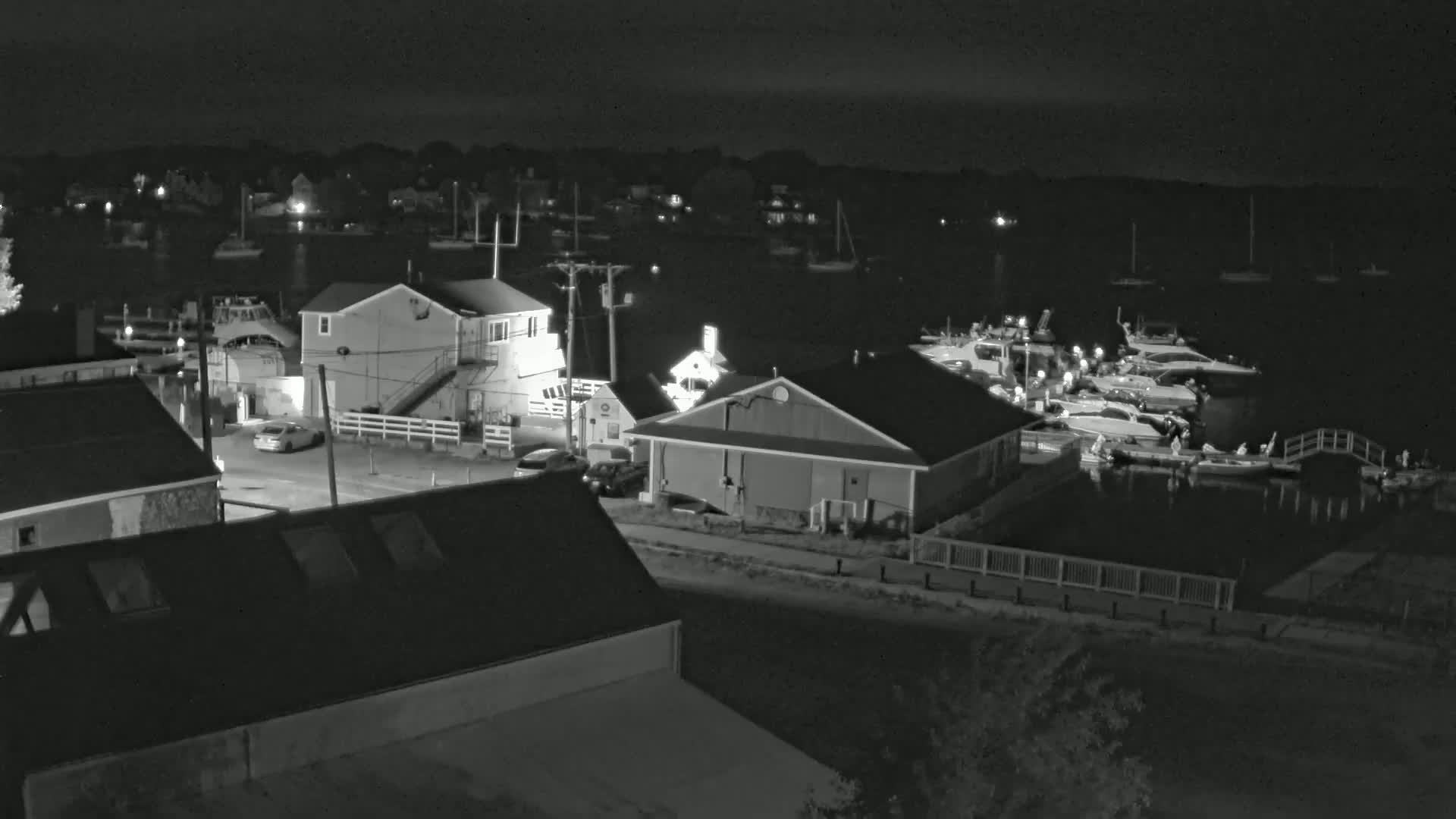 A nighttime view of a harbor with several boats docked, buildings along the shore, and a few cars parked nearby under a clear, dark sky.