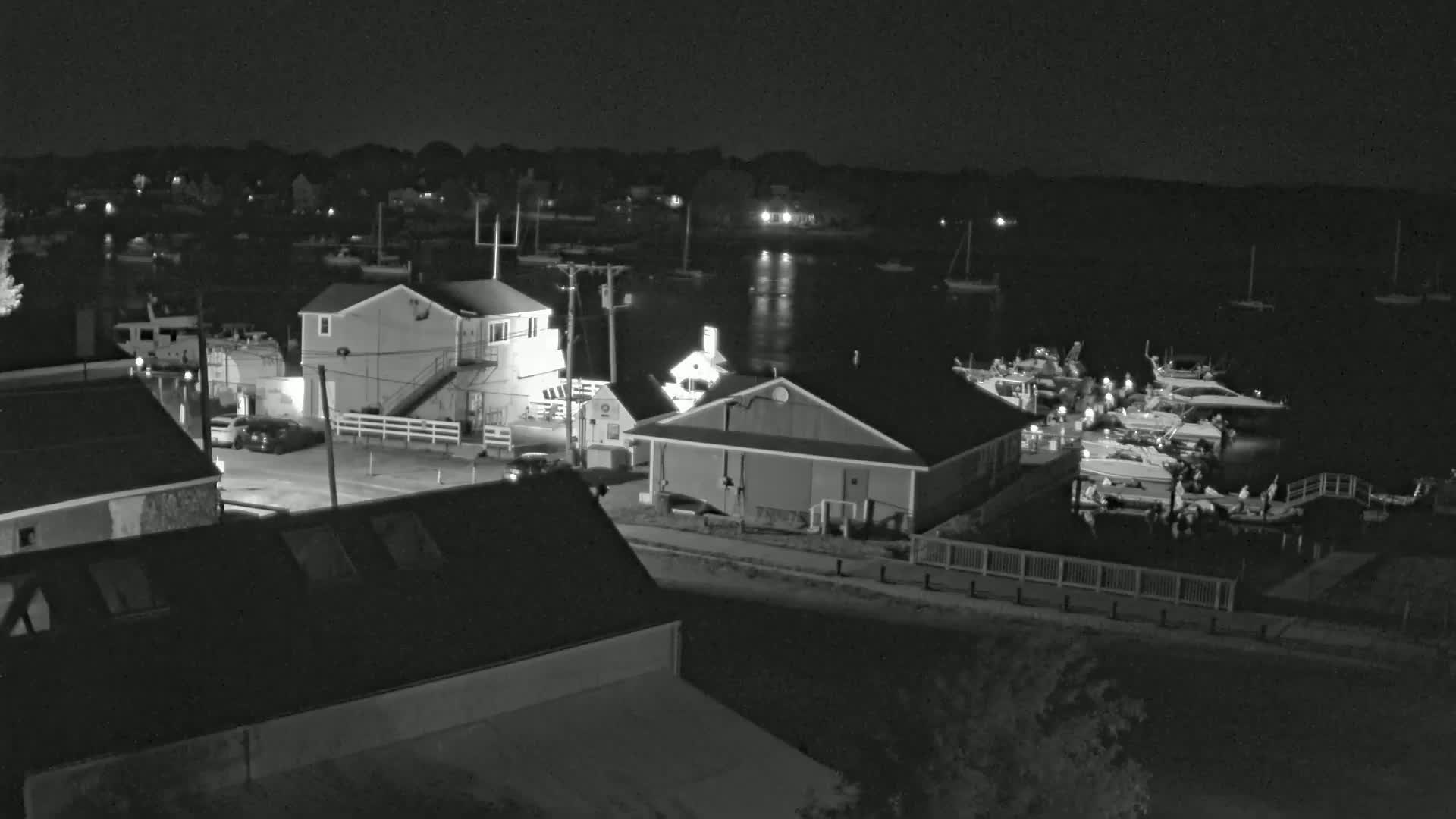A nighttime, black and white view of a harbor with several boats docked, buildings, and cars parked on a roadway next to the water.