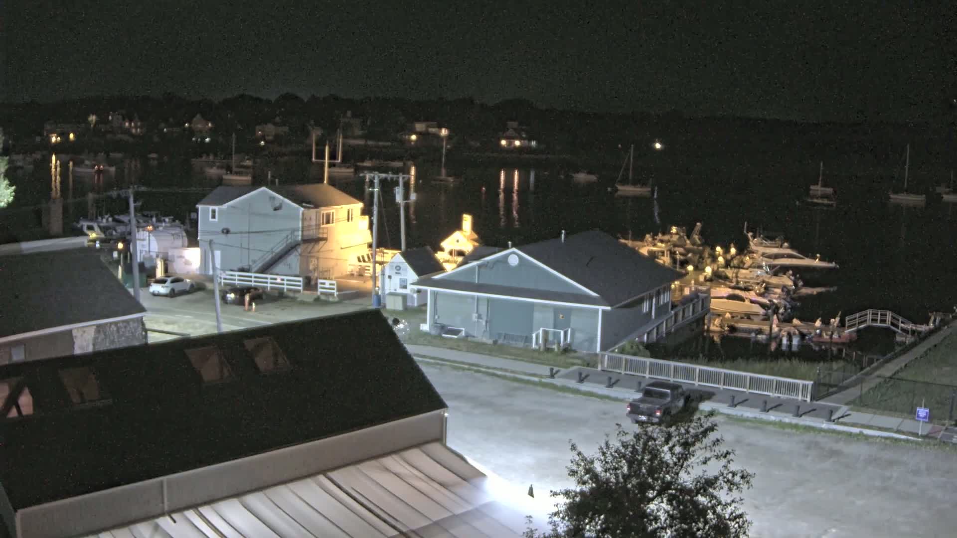 A nighttime, black-and-white view of a waterfront area with several buildings, cars, and numerous boats docked at a marina under a clear sky.