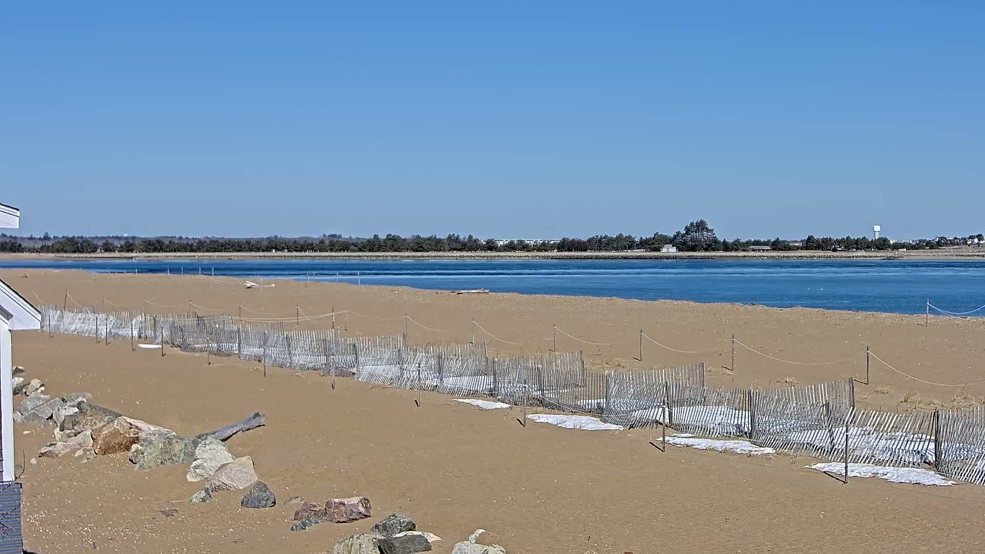 A sandy beach with low wooden fences and white coverings stretches to a calm body of water under a clear blue sky.