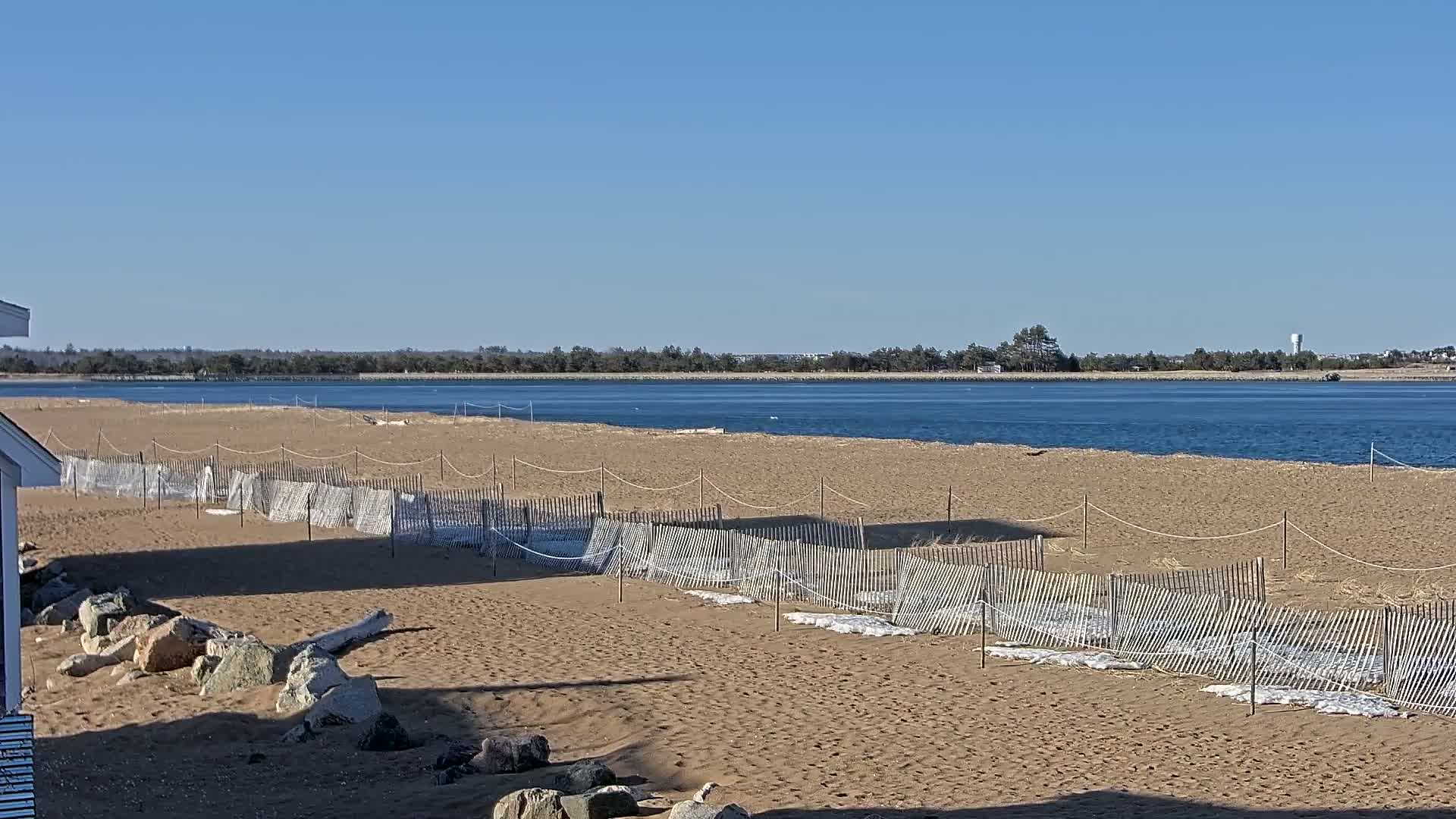 A sandy beach with snow-covered dune fences stretches towards a calm body of water under a clear blue sky.