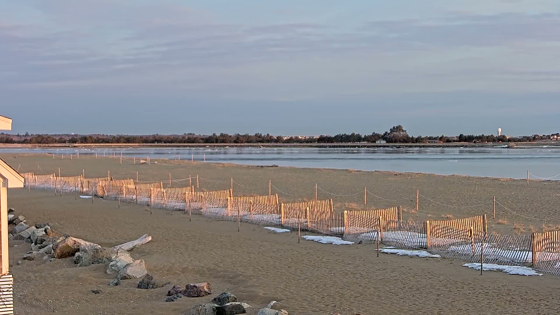 A sandy beach with a low wooden fence partially covered in snow, stretches towards a calm body of water partially frozen under a cloudy sky.