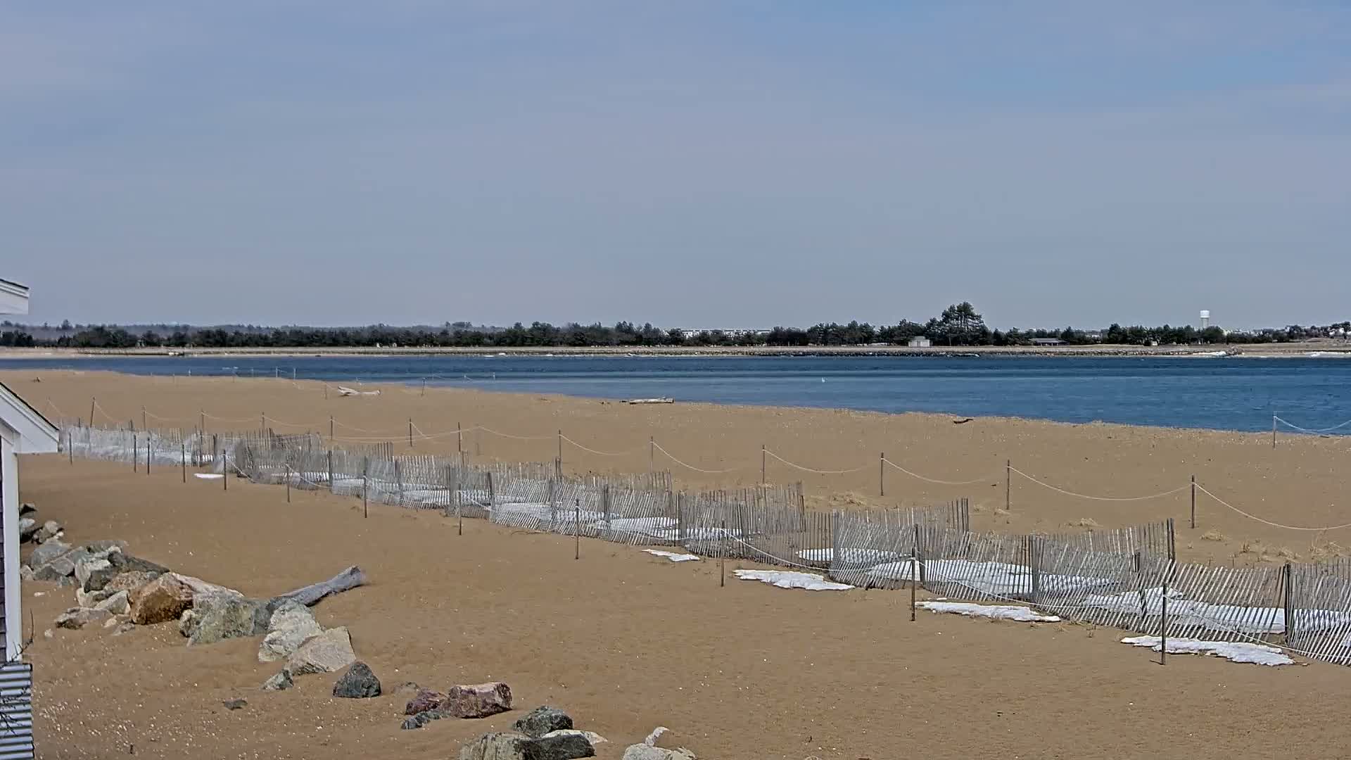A sandy beach with low wooden fences and white coverings stretches toward a calm body of water under a clear, bright sky.