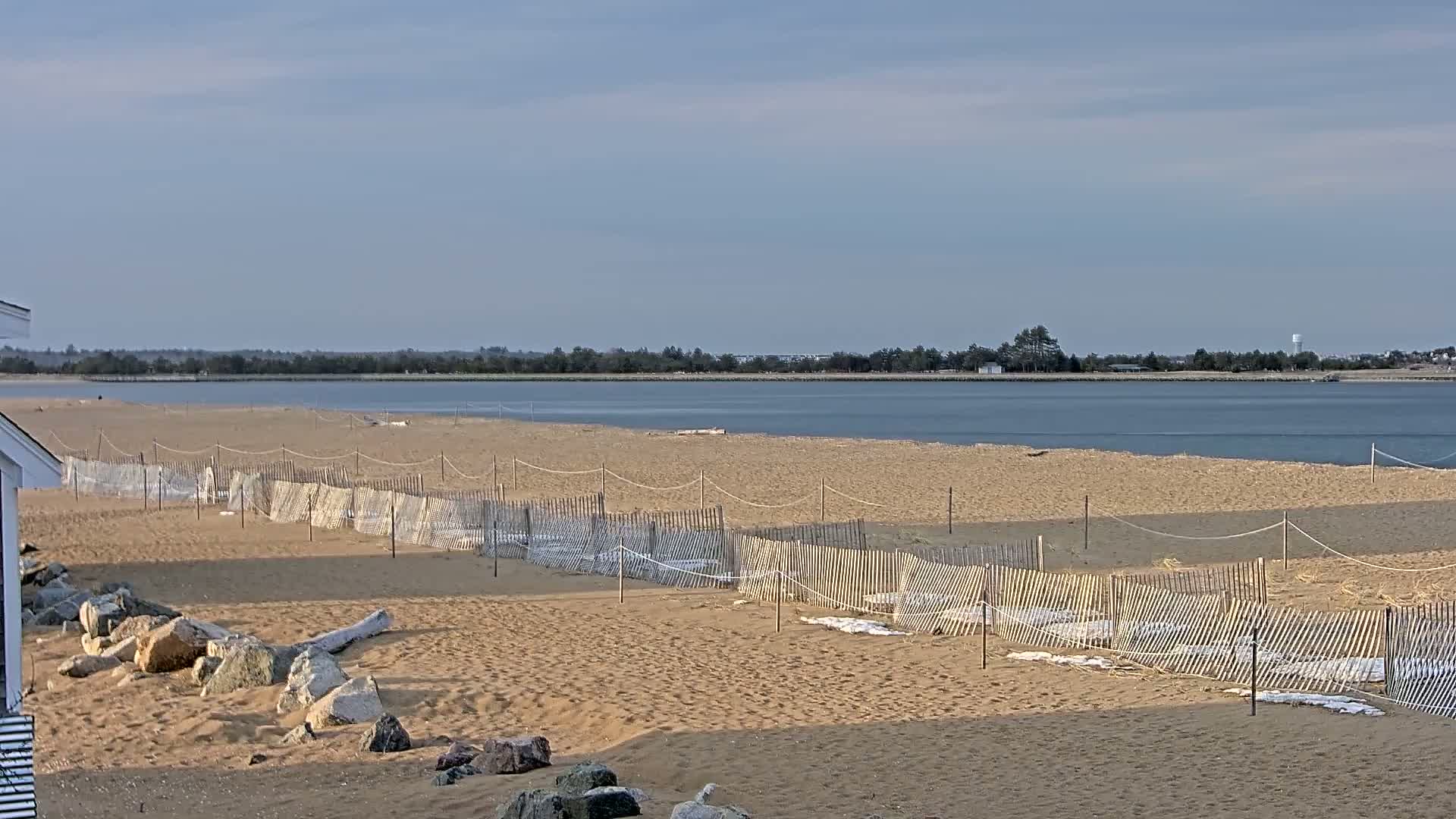 A sandy beach with short wooden fences and white coverings stretches towards a calm body of water under a clear sky.