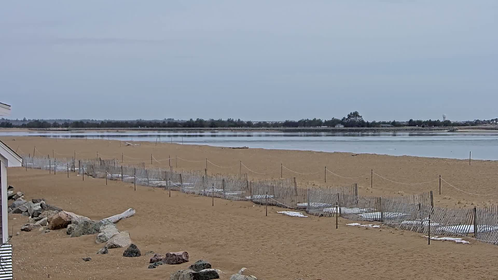 A sandy beach with a low wooden fence and partially frozen water stretches to a tree-lined horizon under a cloudy sky.