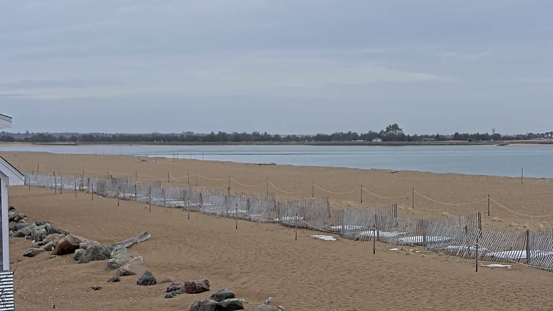 A sandy beach with a low wooden fence stretches along a calm body of water under an overcast sky.