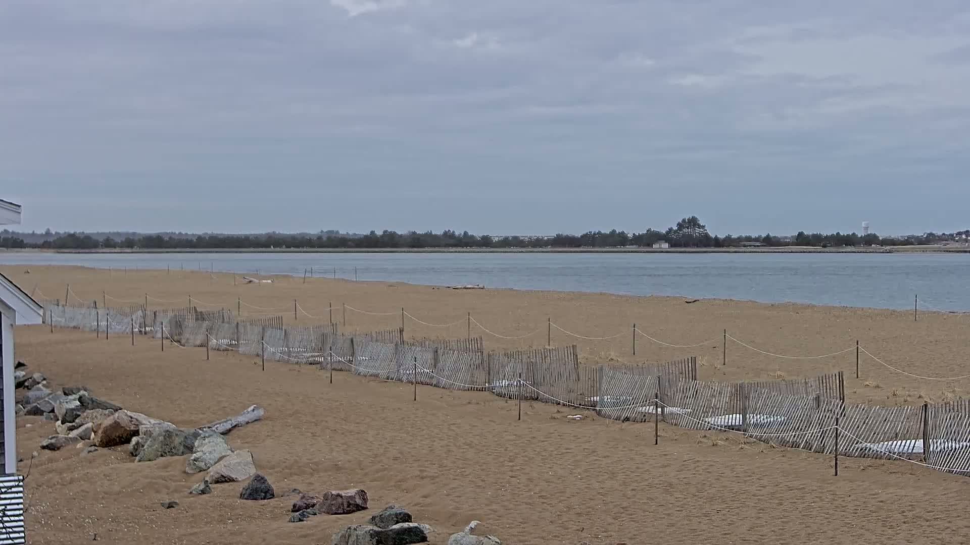 A sandy beach with a low wooden fence stretches along a calm body of water under an overcast sky.