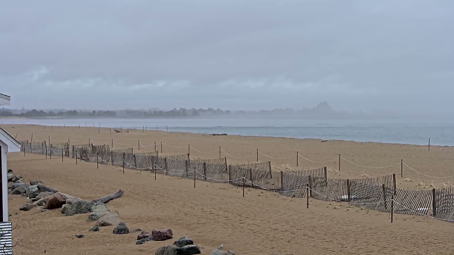 A sandy beach with a low wooden fence stretches towards a calm, hazy ocean under an overcast sky.