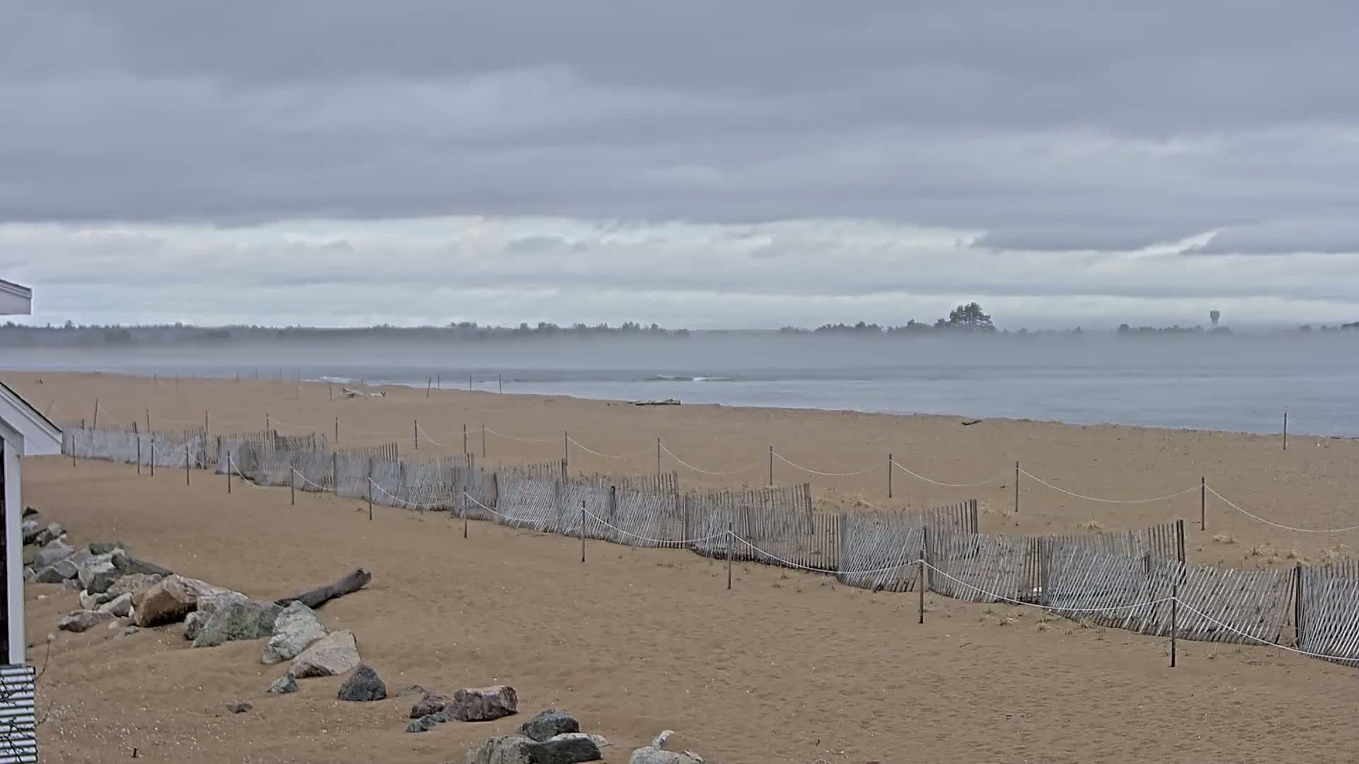 A sandy beach with a low wooden fence stretches towards a calm, foggy body of water under a cloudy sky.