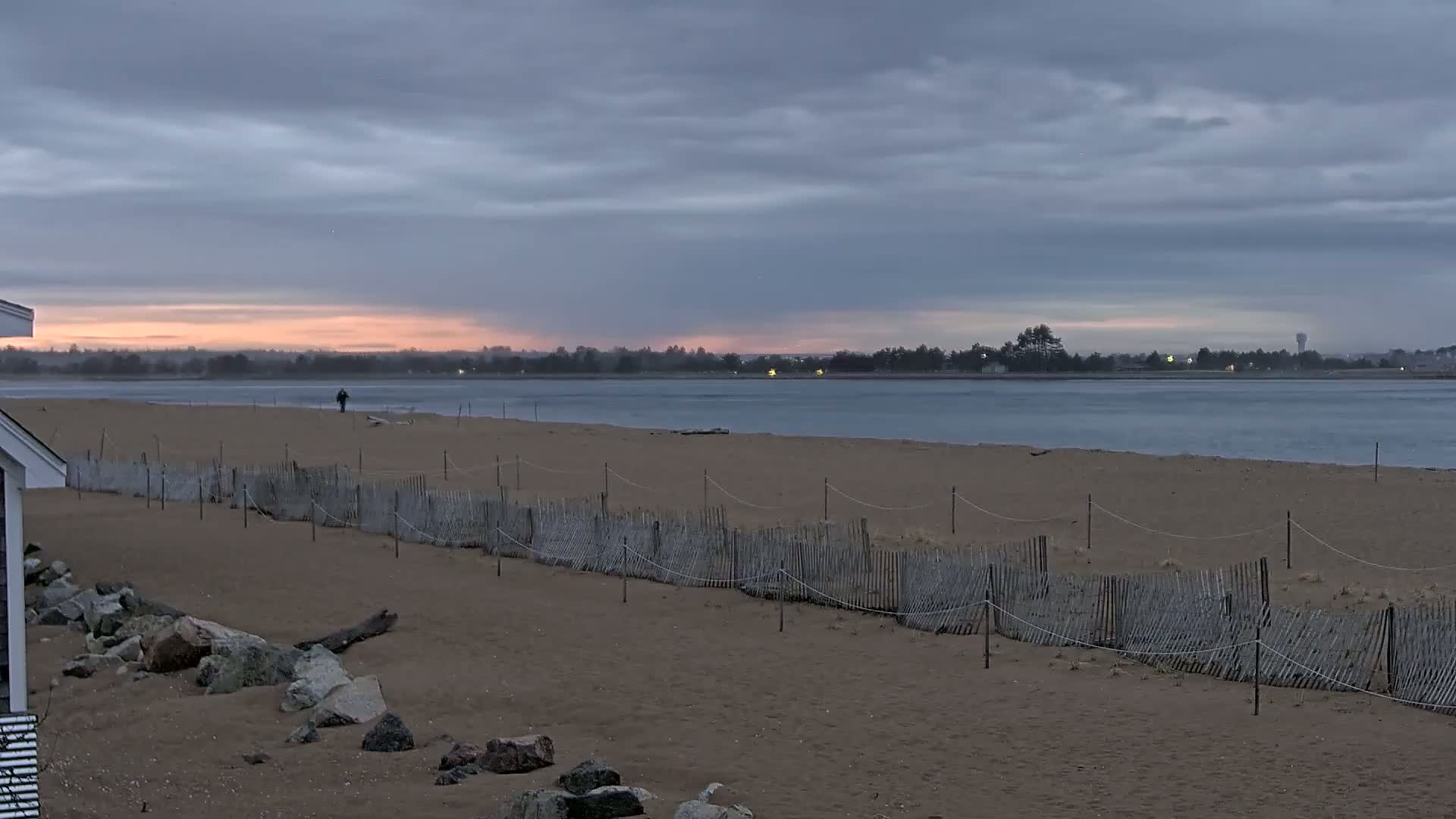 A sandy beach with a low wooden fence stretches along a calm body of water under a cloudy, sunrise sky.