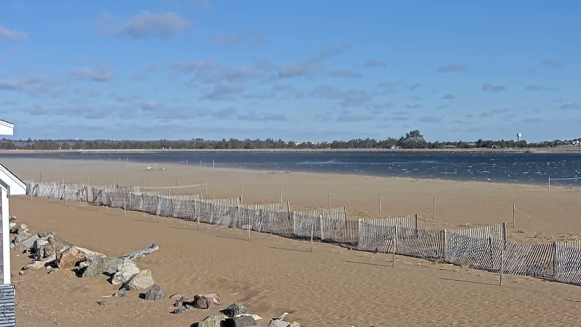 A sandy beach with a wooden fence stretches along a calm, wavy body of water under a mostly sunny sky.