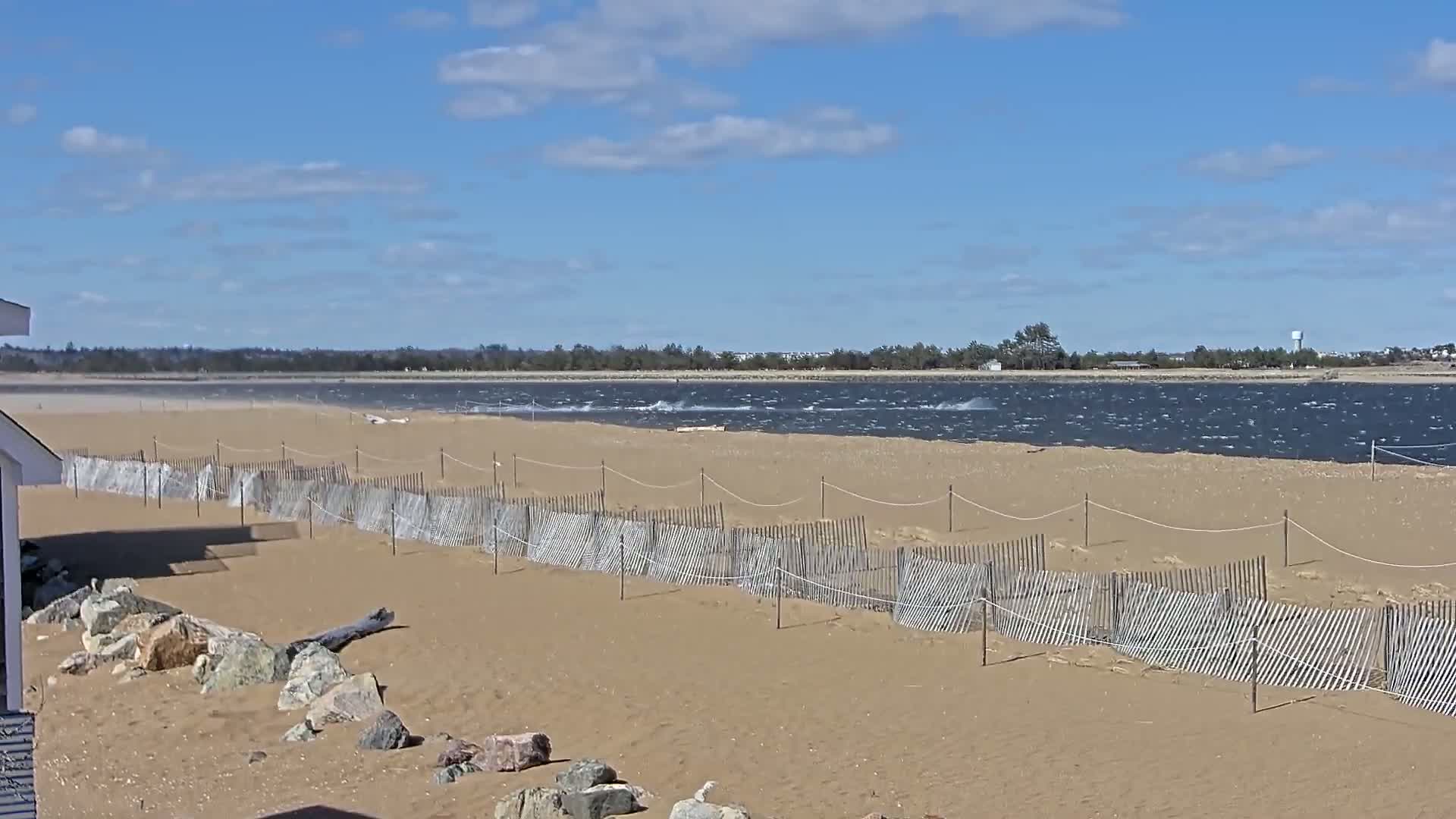 A sandy beach with a low wooden fence stretches along a windy, choppy body of water under a partly cloudy blue sky.