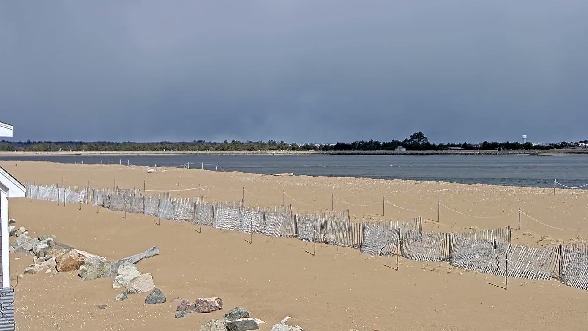 A sandy beach with a low wooden fence stretches towards a calm body of water under a mostly cloudy sky.