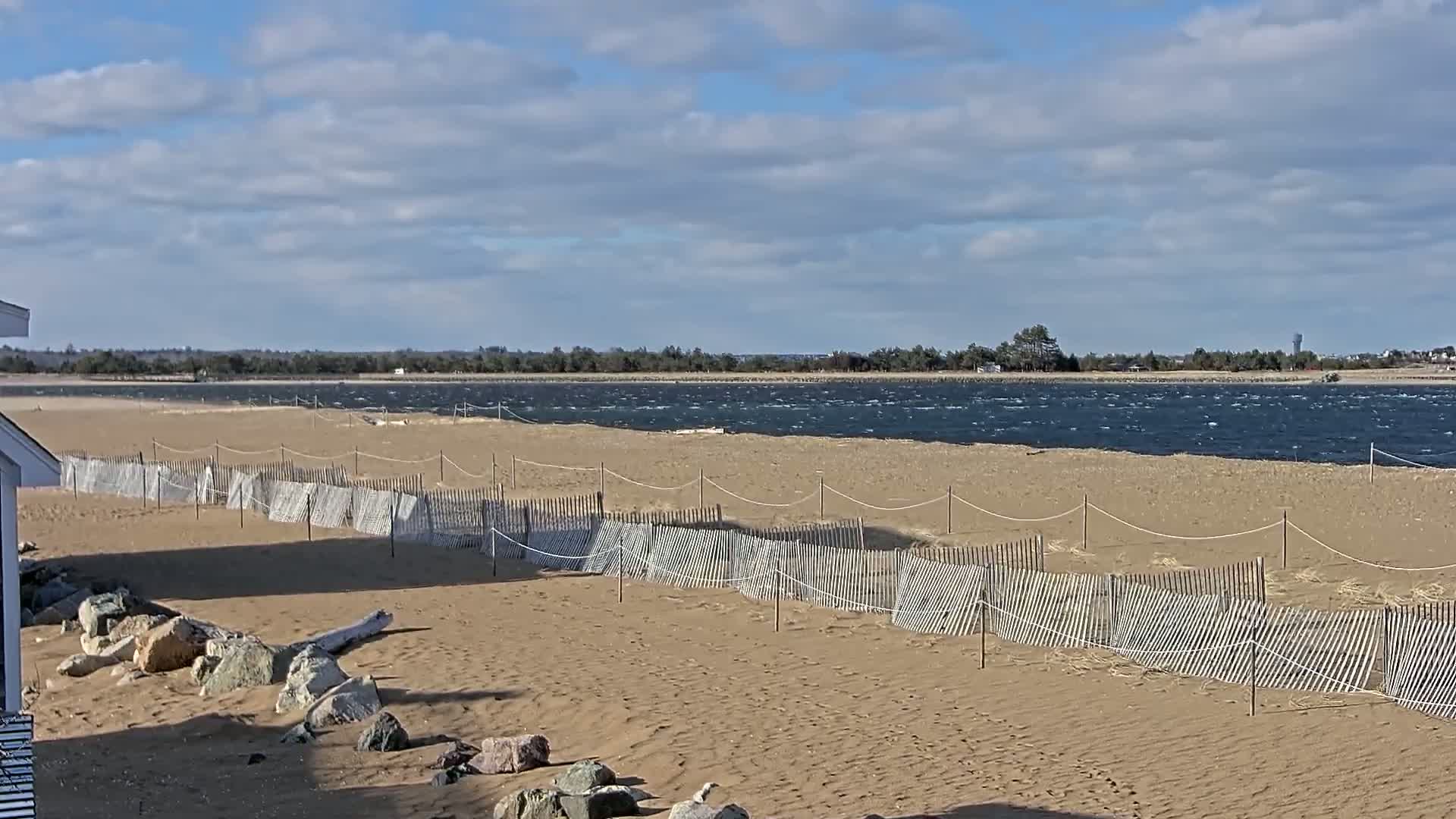 A sandy beach with a low, damaged wooden fence stretches towards a choppy body of water under a partly cloudy sky.