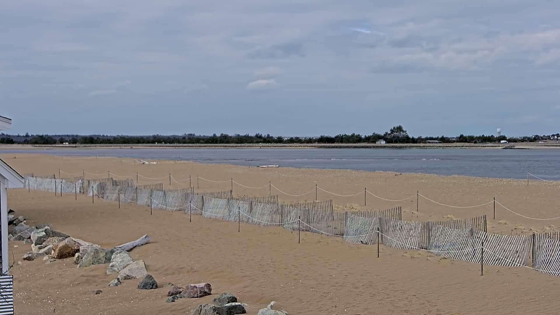 A sandy beach with a low wooden fence stretches towards a calm body of water under a cloudy sky.