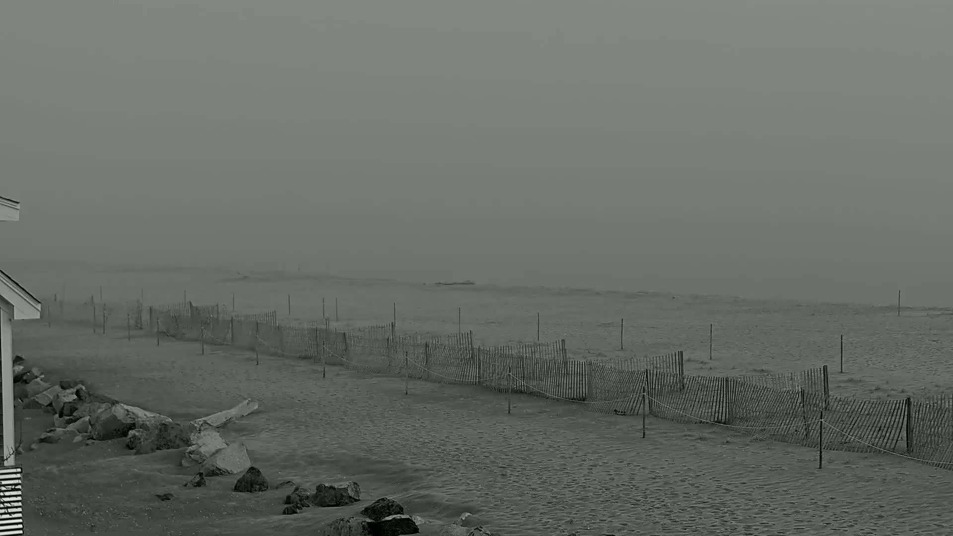 A sandy beach stretches to a hazy ocean under a foggy sky,  partially obscured by a weathered fence running along the shore.