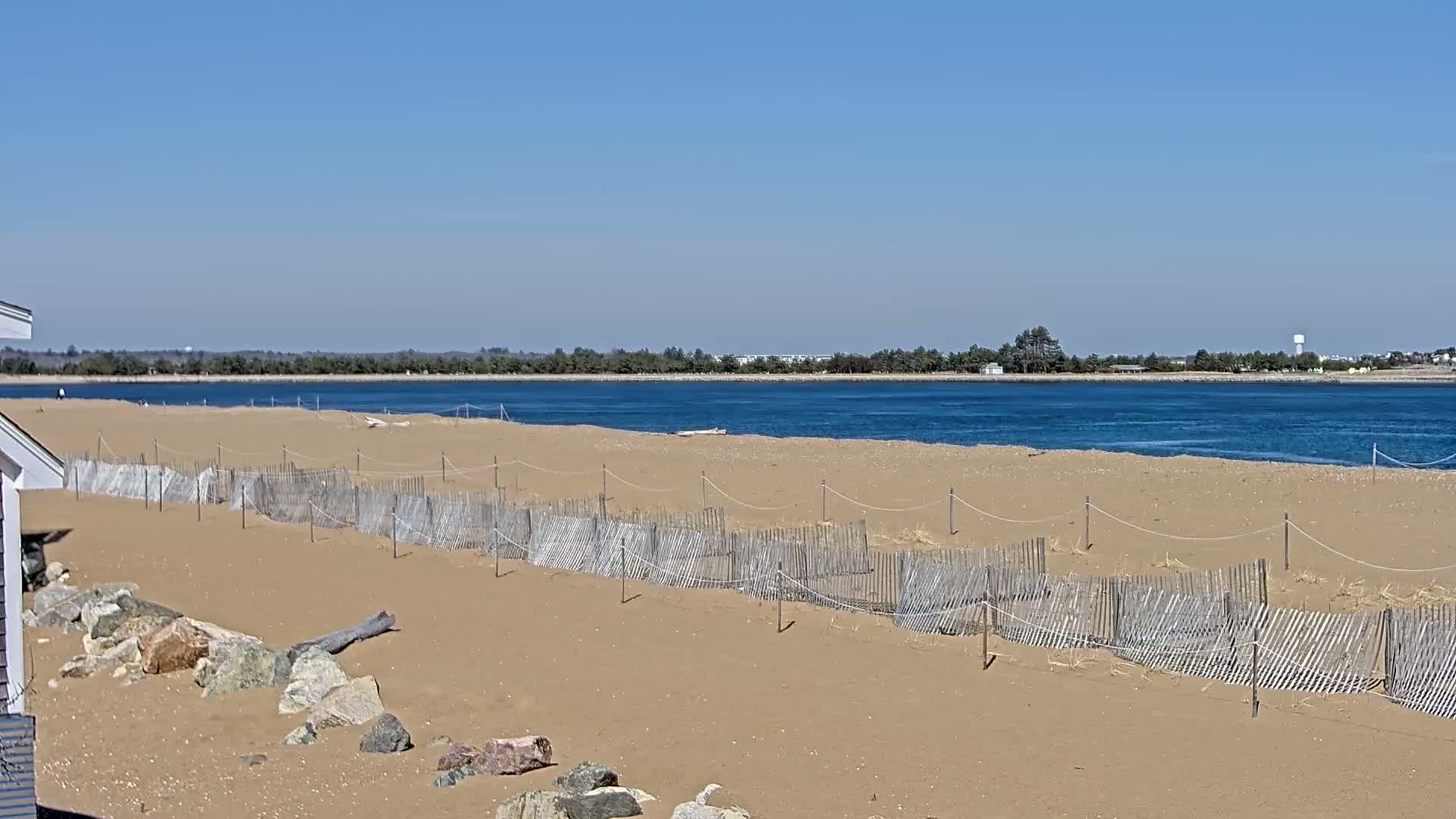 A sandy beach with short, wooden fences stretches along a calm body of water under a clear blue sky.
