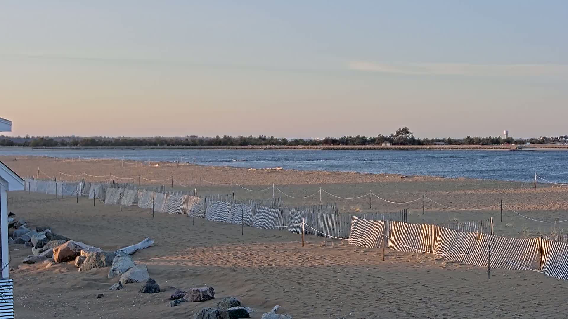 A sandy beach with a low wooden fence stretches towards a calm body of water under a clear, light-blue sky.