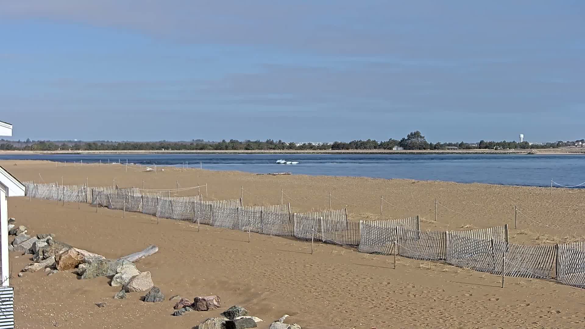 A sandy beach with a short wooden fence stretches toward a calm body of water under a clear blue sky.