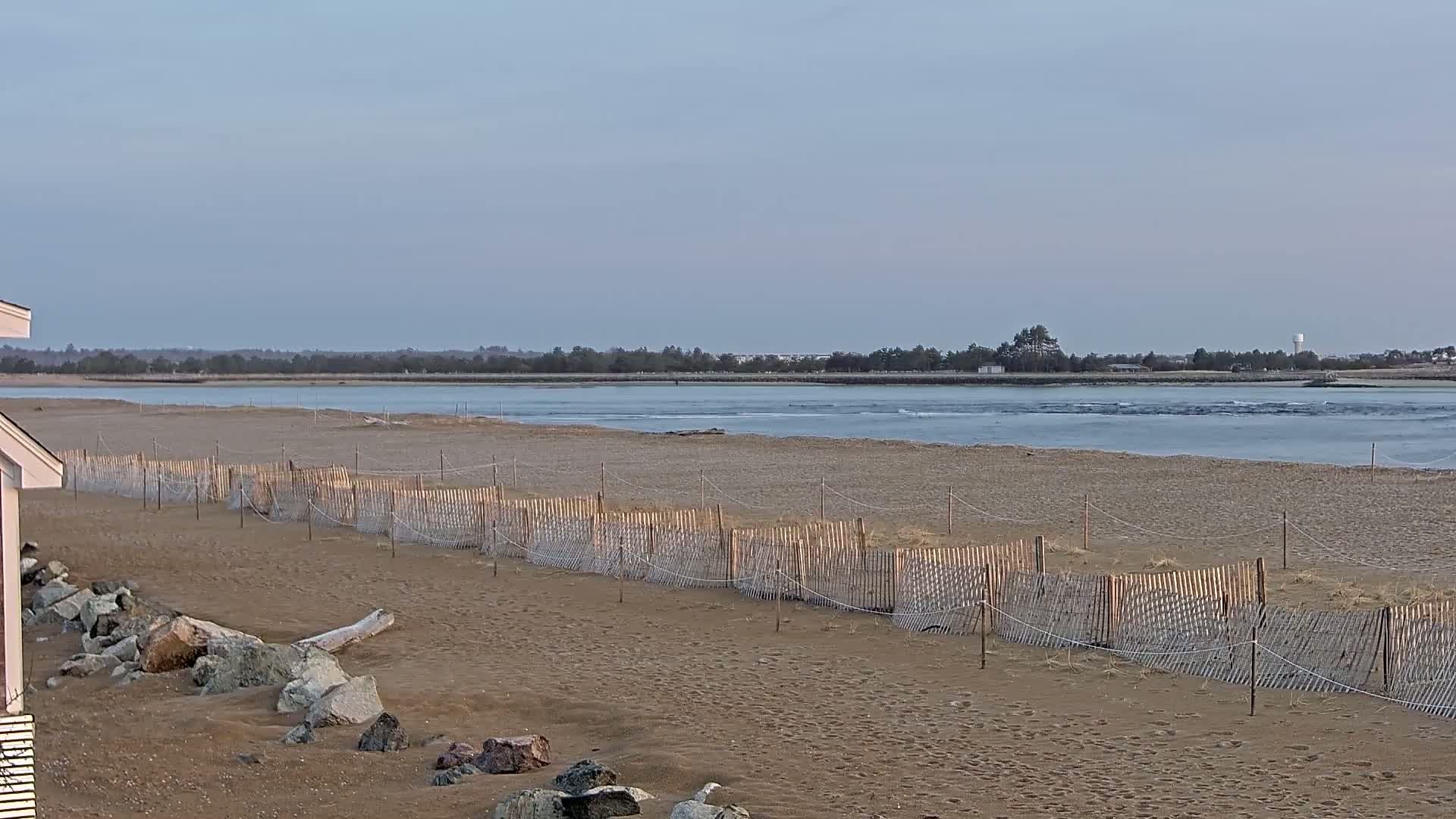 A sandy beach with a low wooden fence stretches toward a calm body of water under a clear, pale sky.
