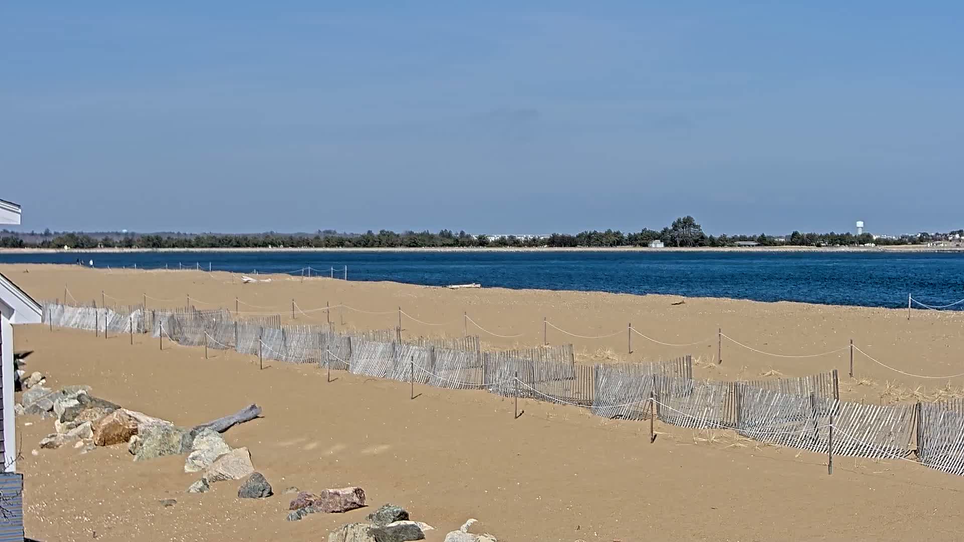 A sandy beach with a low wooden fence stretches along a calm body of blue water under a clear blue sky.