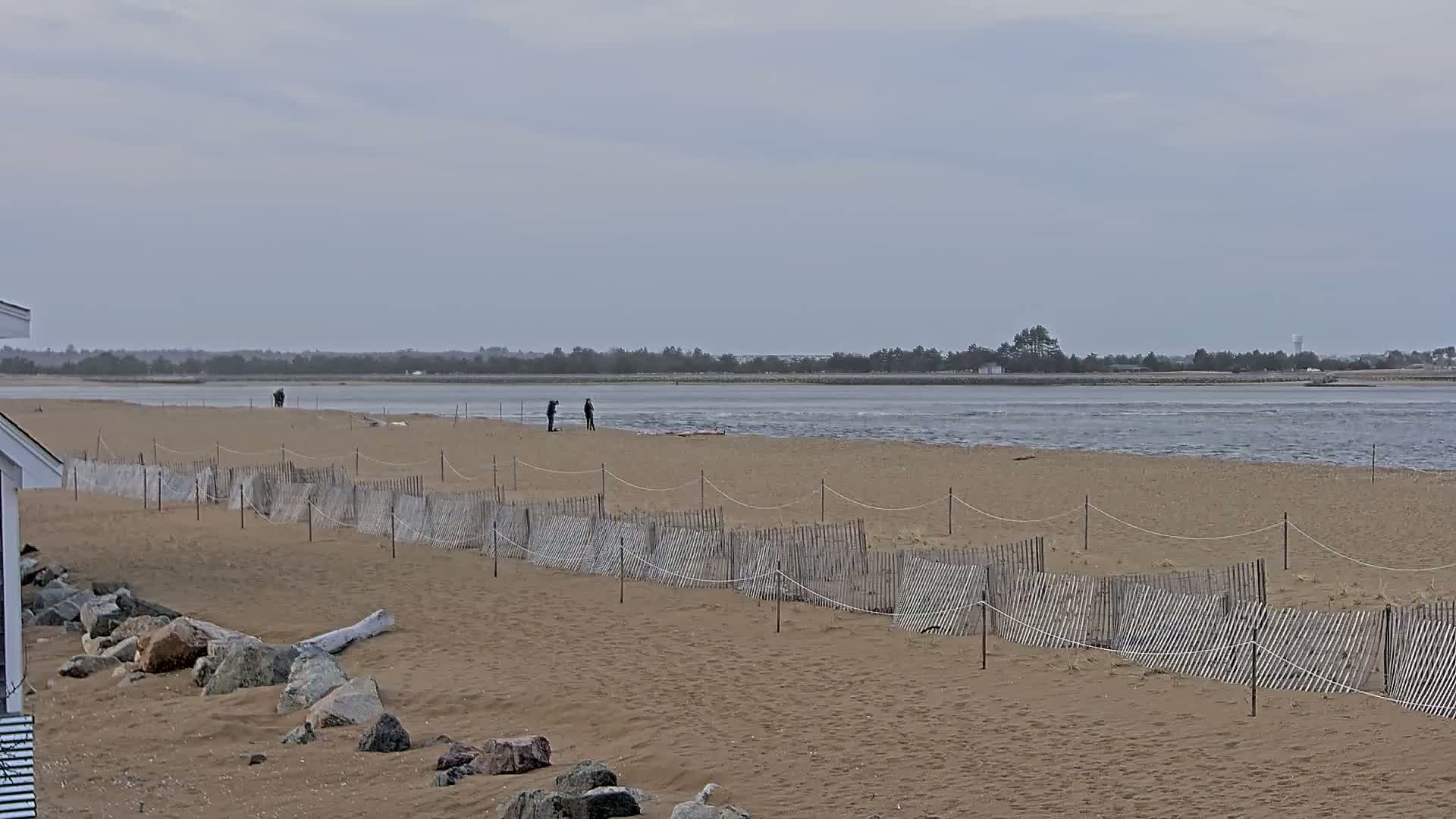 A sandy beach with a low wooden fence stretches toward a calm, gray body of water under an overcast sky, and a few people are standing on the beach.