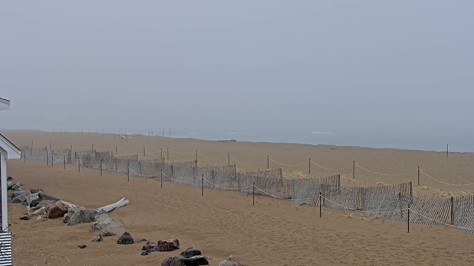 A sandy beach stretches to a foggy ocean, with a wooden fence running parallel to the shore, and a rocky area near a building.