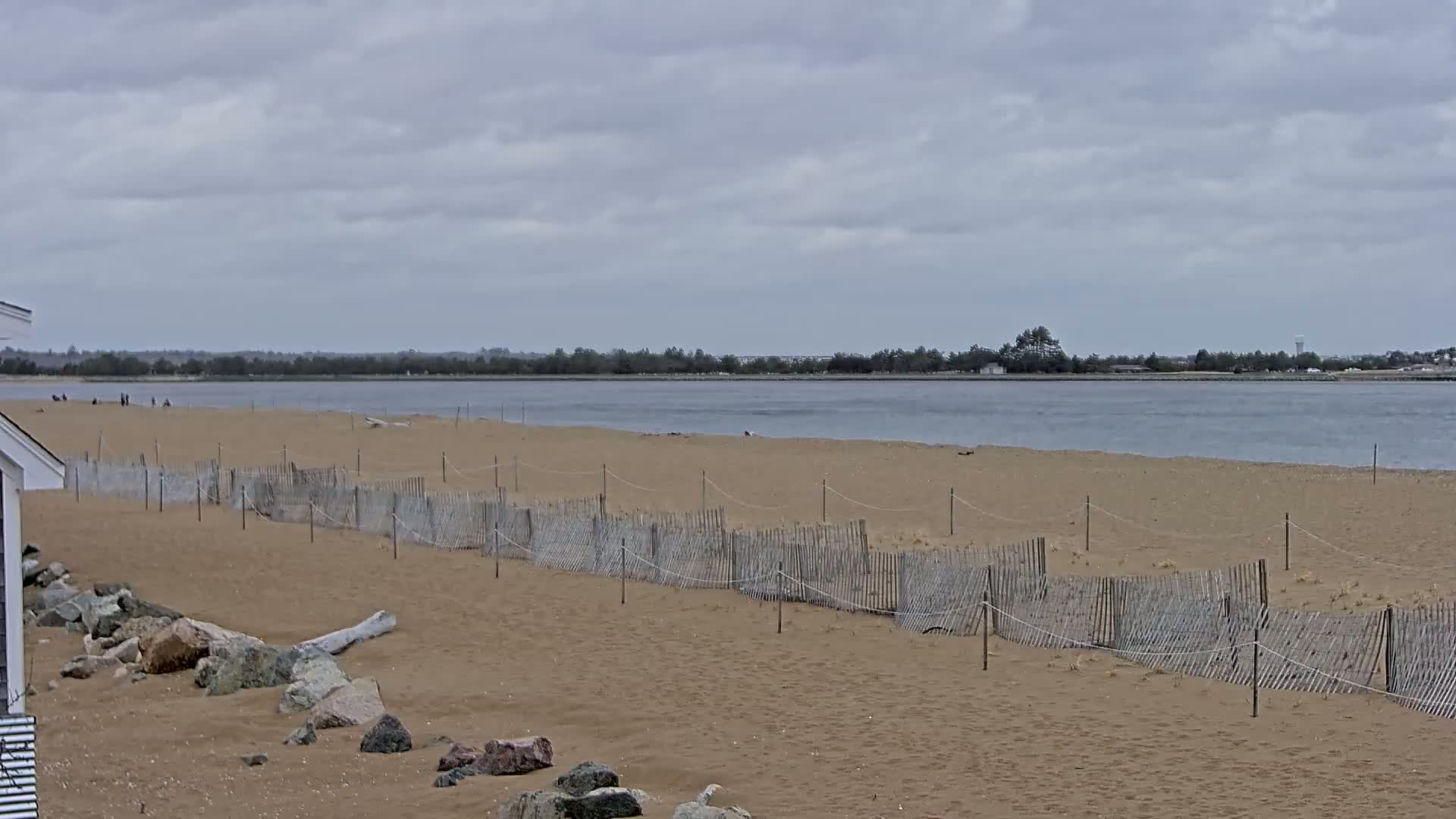 A sandy beach with a low wooden fence stretches along a calm body of water under a cloudy sky.