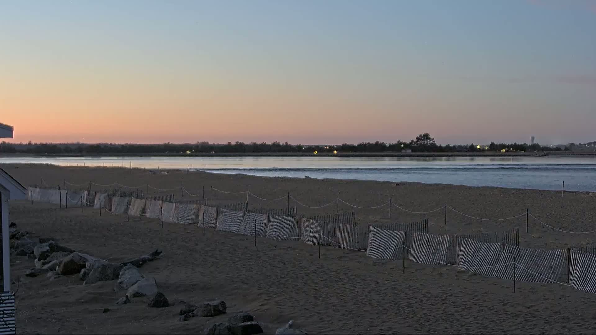 A sandy beach with a low wooden fence stretches toward a calm body of water under a pinkish sunset sky.