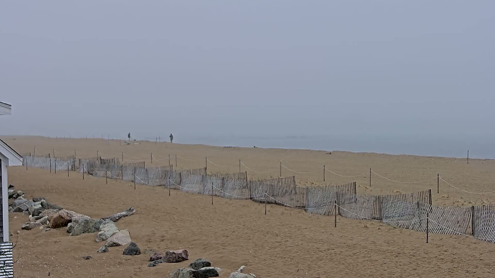 A foggy beach scene shows a long, low wooden fence running parallel to the shoreline, with a few people visible in the distance on the sand.