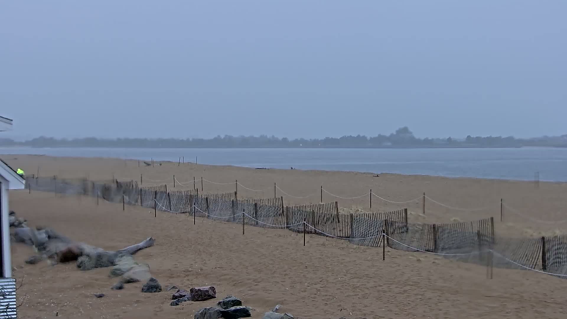 A sandy beach with a wooden fence stretches along a calm, gray body of water under an overcast sky.