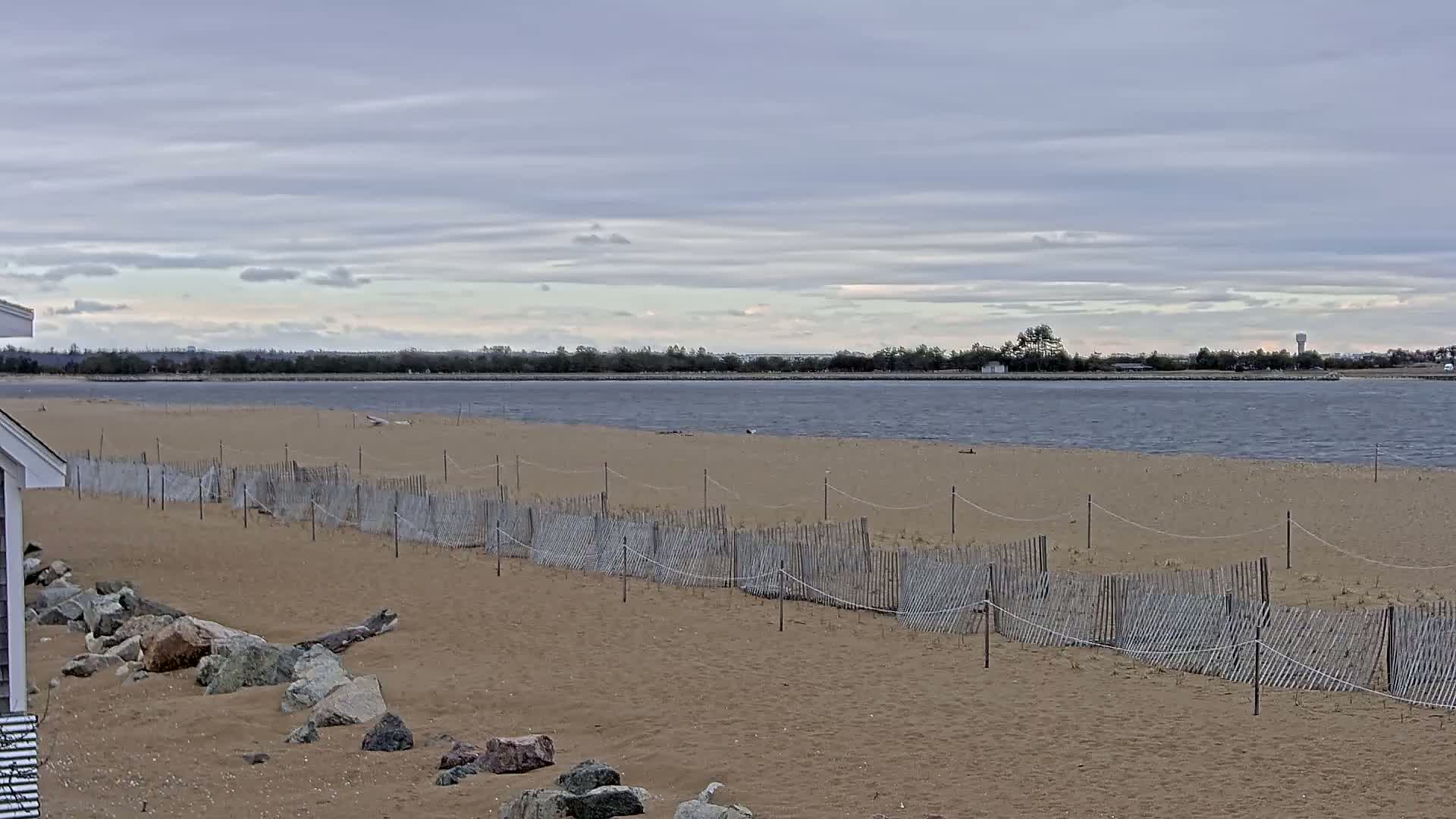 A sandy beach with a low wooden fence stretches along a calm body of water under a cloudy sky.