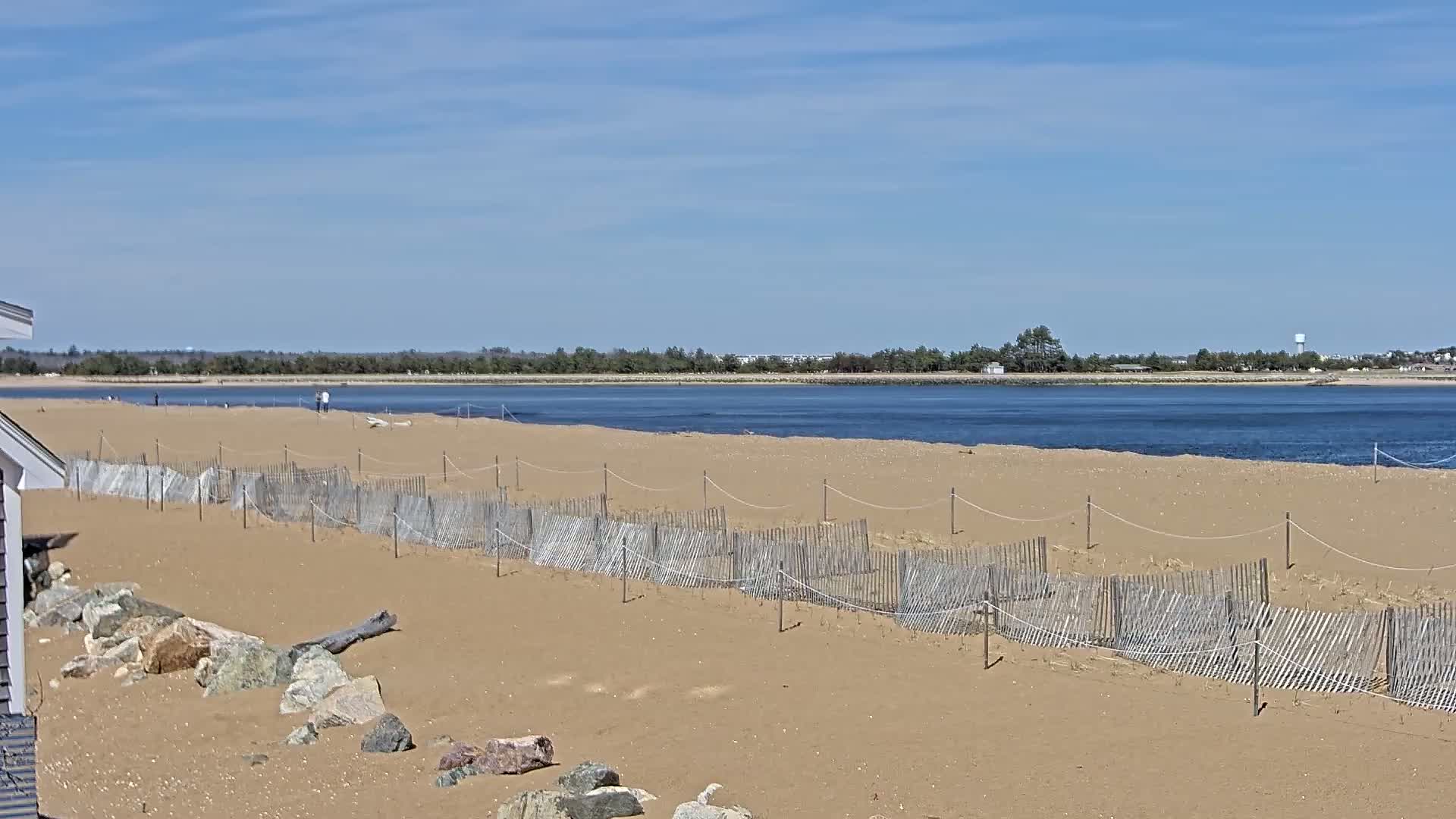 A sandy beach with a low wooden fence stretches along a calm body of water under a clear blue sky.
