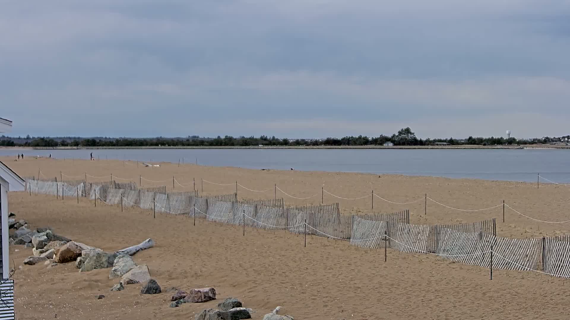 A sandy beach with a low wooden fence stretches along calm, grey water under a cloudy sky.