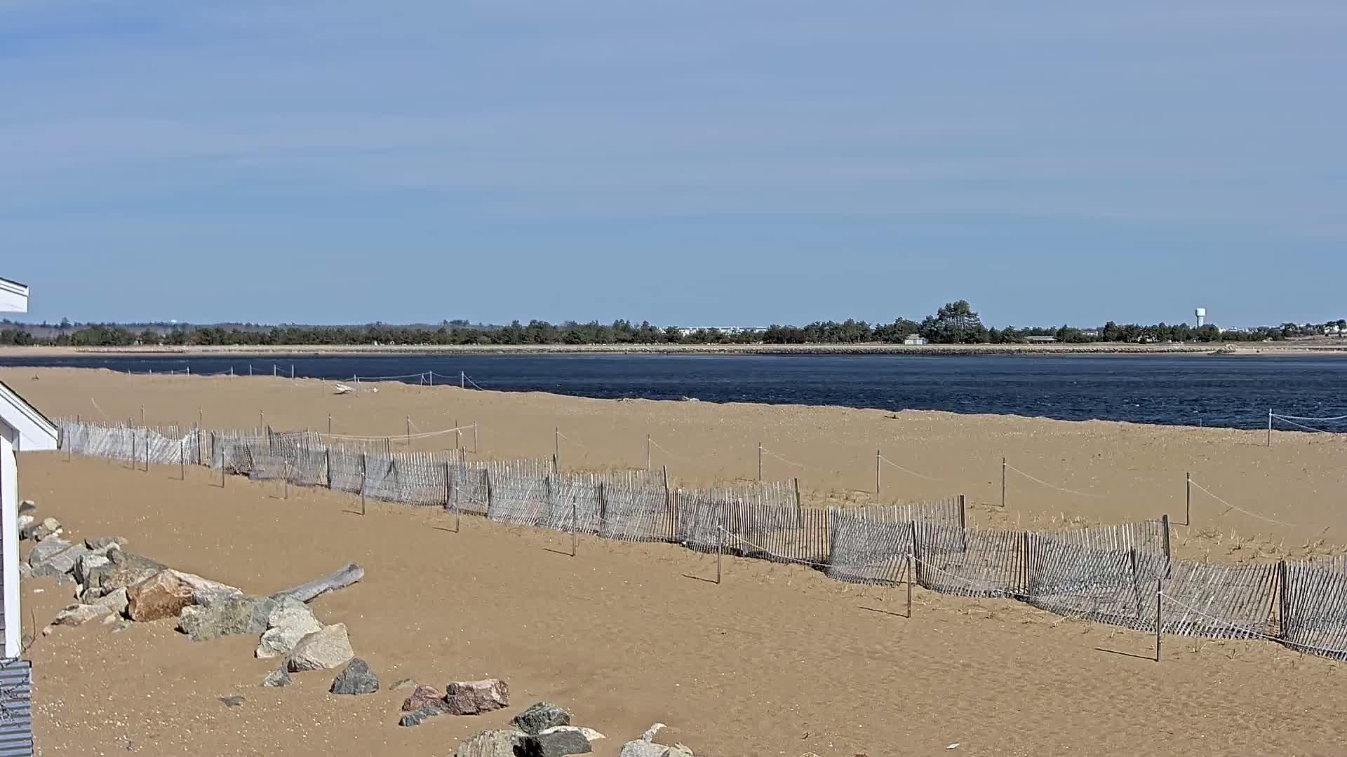 A sandy beach with a low wooden fence stretches to a calm, dark-colored body of water under a clear blue sky.