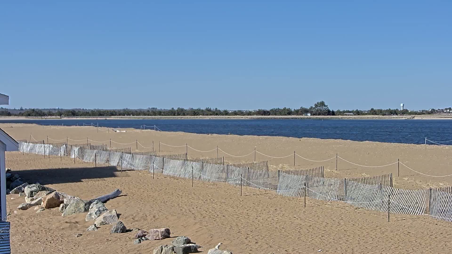 A sandy beach with a low wooden fence stretches along a calm body of water under a clear blue sky.