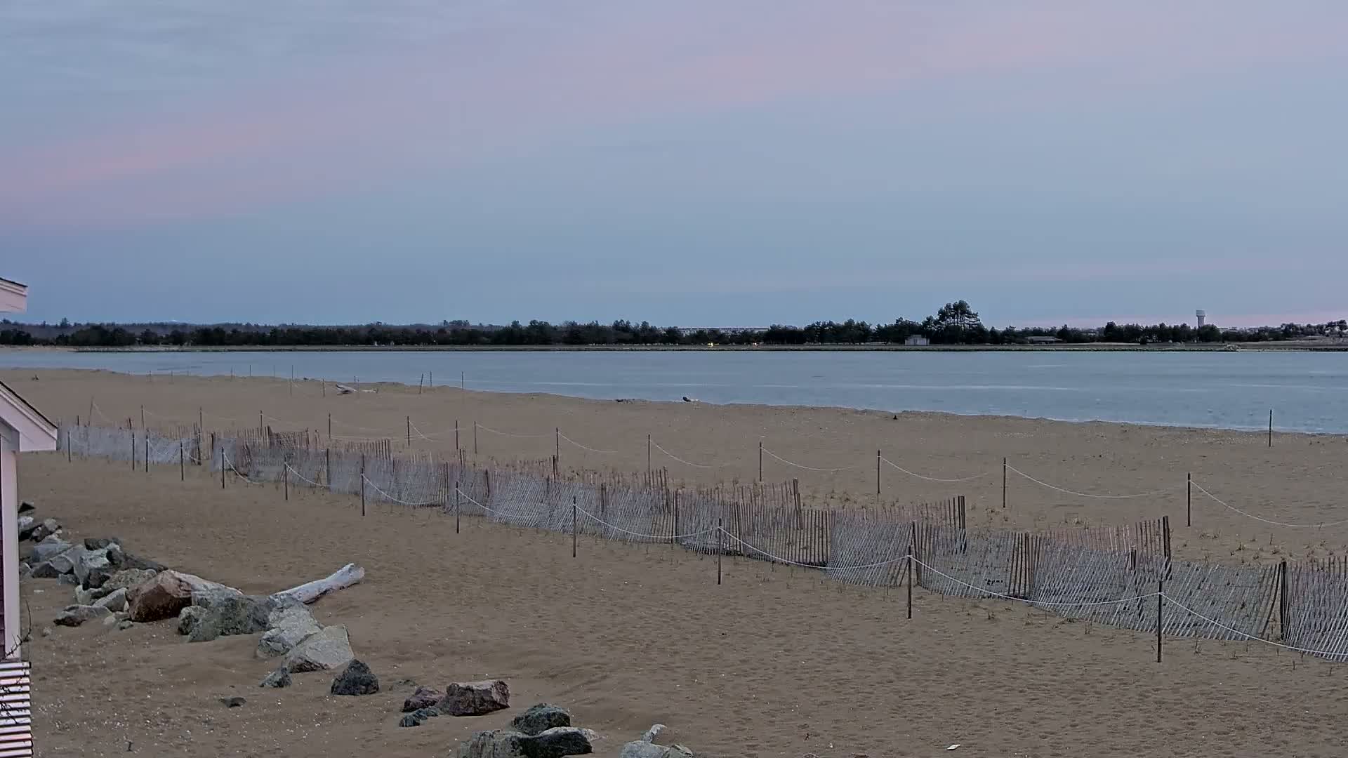 A sandy beach with a low wooden fence stretches along a calm body of water under a pale, overcast sky.