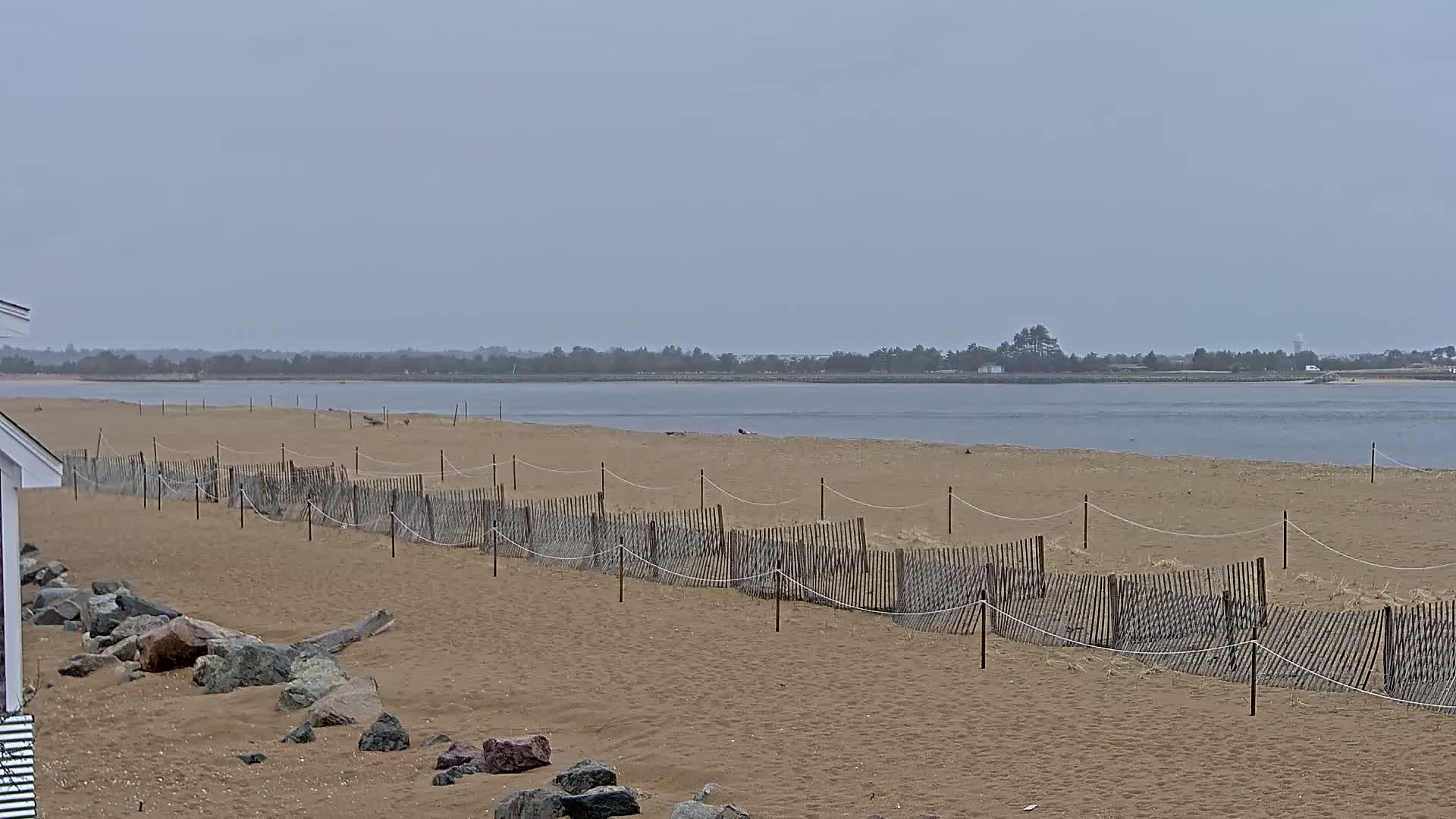 A sandy beach with a low wooden fence stretches along a calm, grey body of water under an overcast sky.