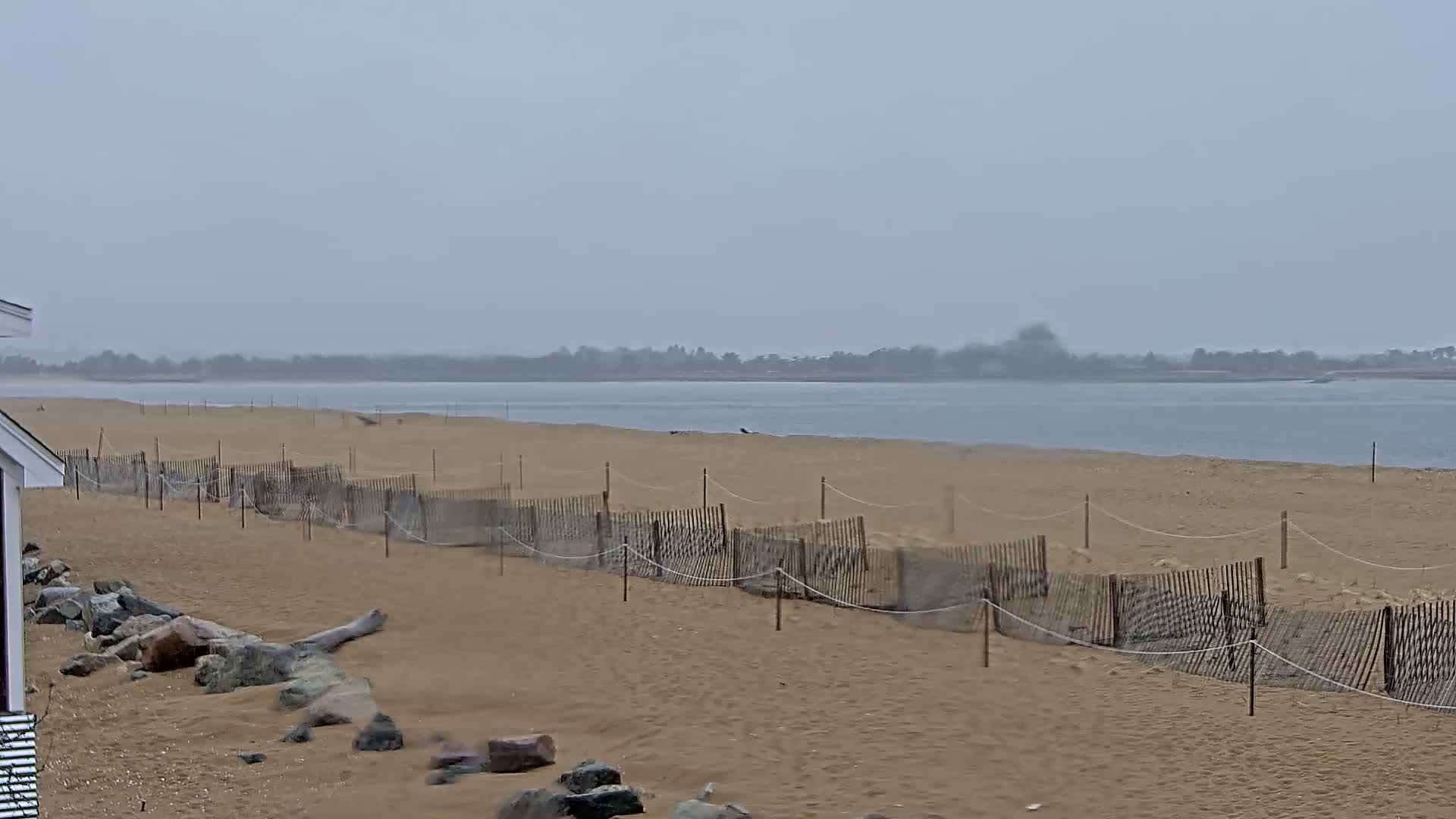 A sandy beach with a low wooden fence stretches along a calm body of water under an overcast sky.