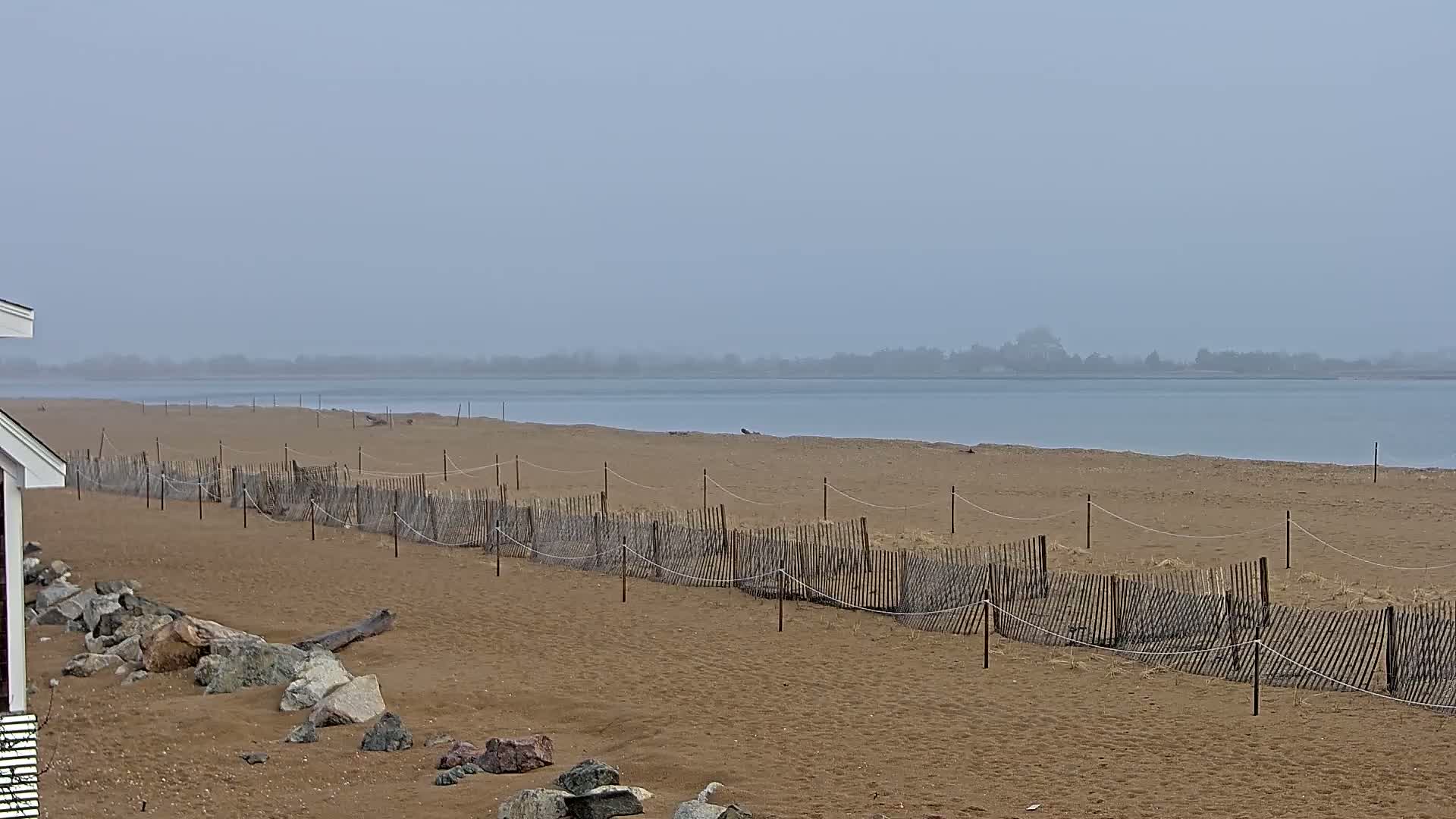 A sandy beach with a low wooden fence stretches along a calm, foggy body of water, backed by a hazy shoreline.