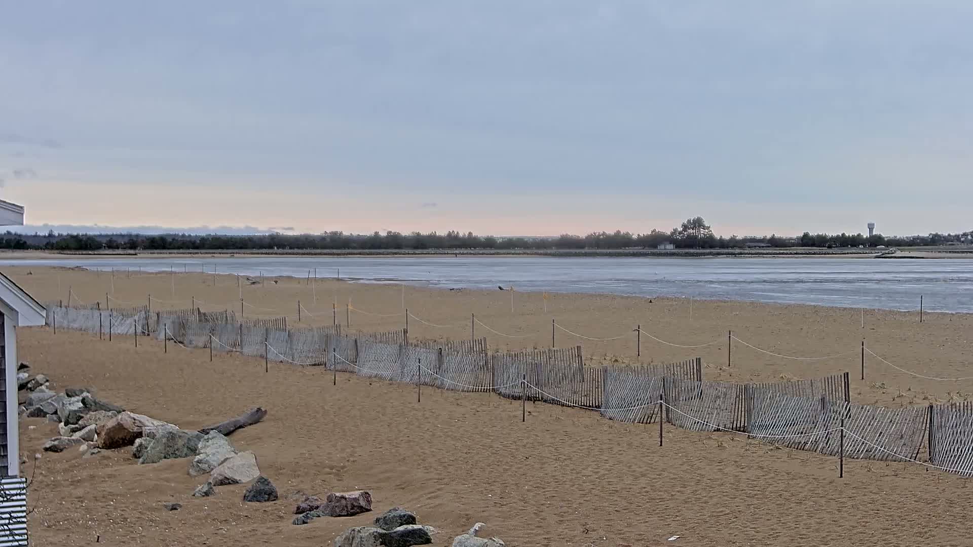 A sandy beach with a low wooden fence stretches towards a calm, partly icy body of water under an overcast sky.