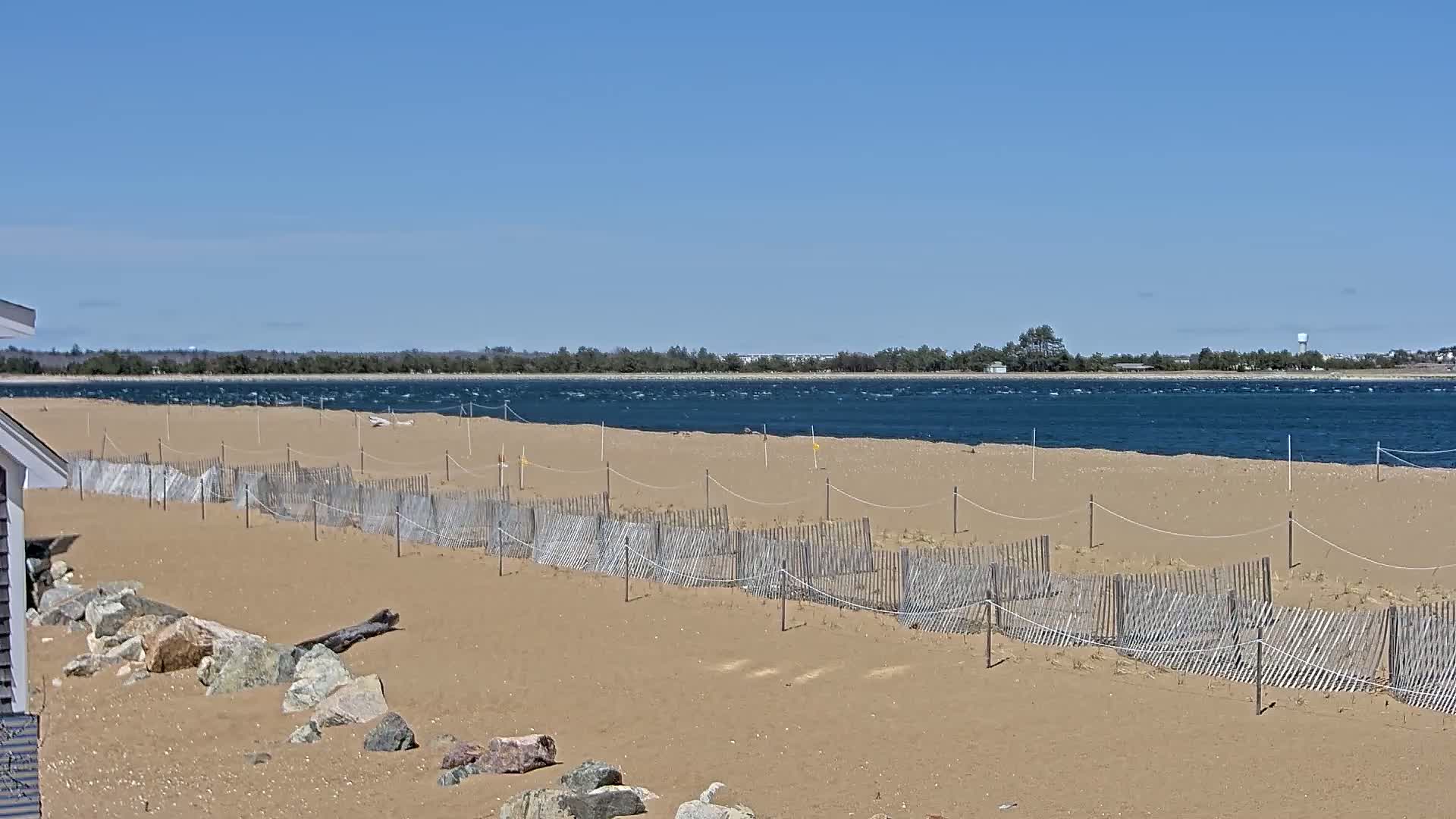 A sandy beach with a low wooden fence stretches along a windy, dark-blue body of water under a clear blue sky.