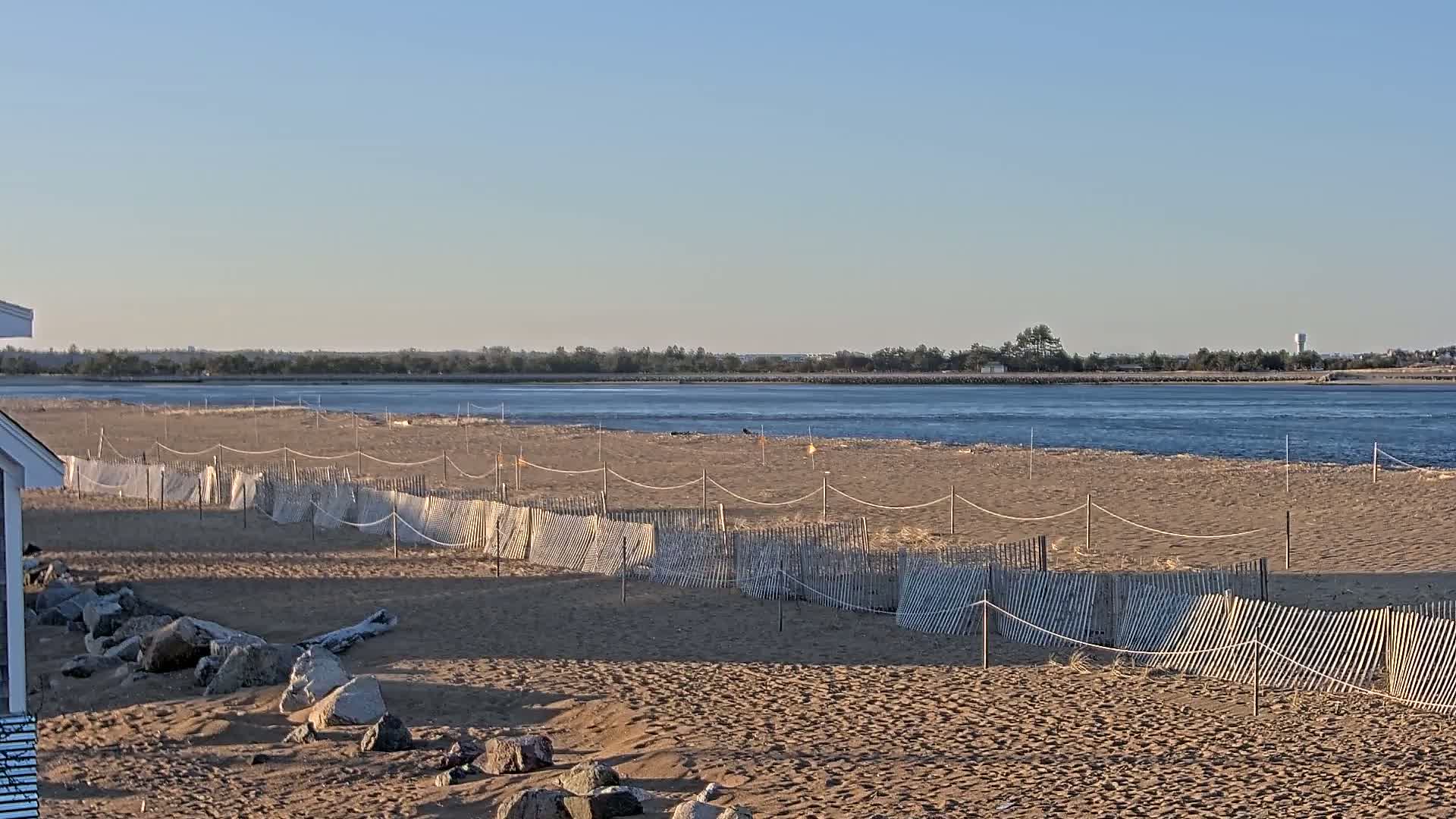 A sandy beach with a low wooden fence separating it from a calm body of water under a clear, sunny sky.