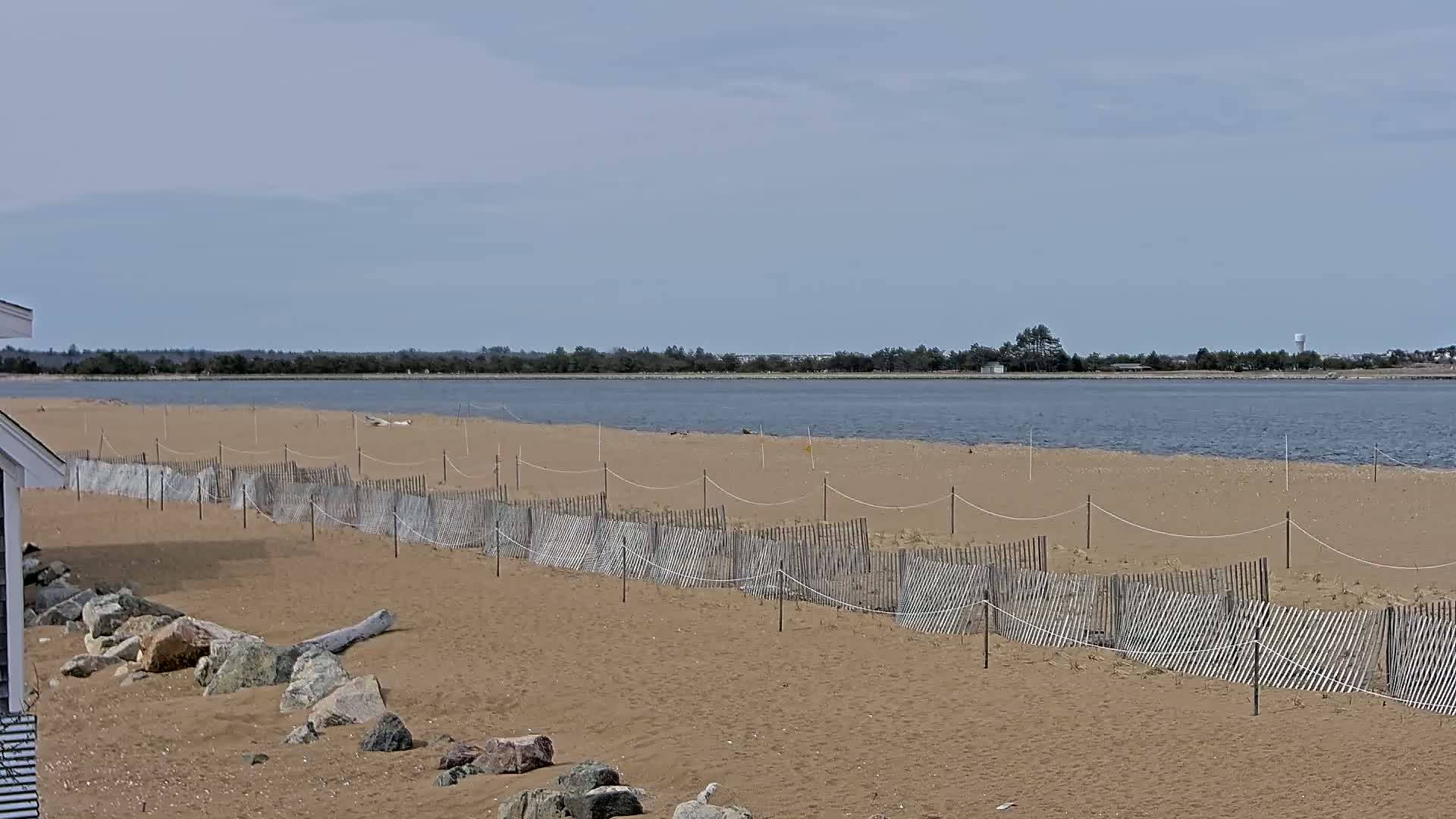 A sandy beach with a low wooden fence stretches along a calm body of water under a cloudy sky.