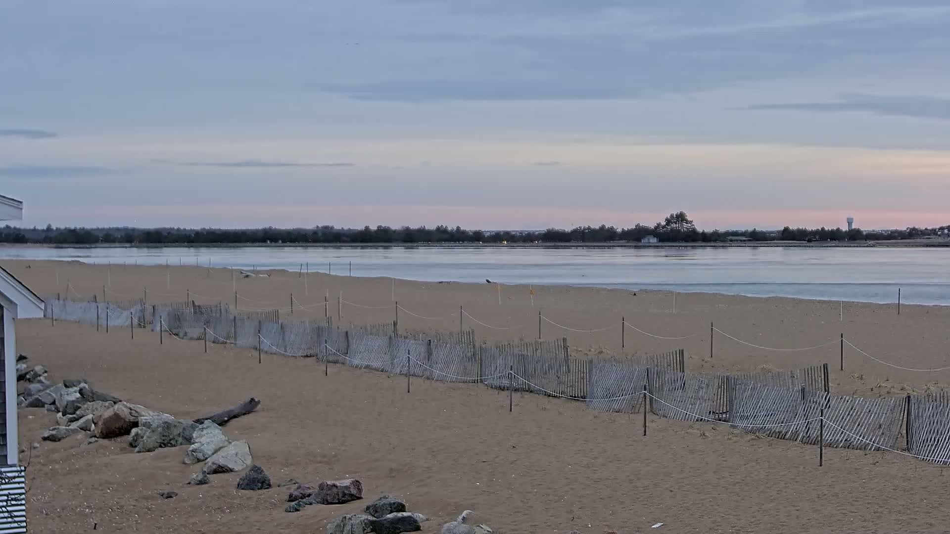 A sandy beach with a low wooden fence stretches towards a calm body of water under a pale, cloudy sky.